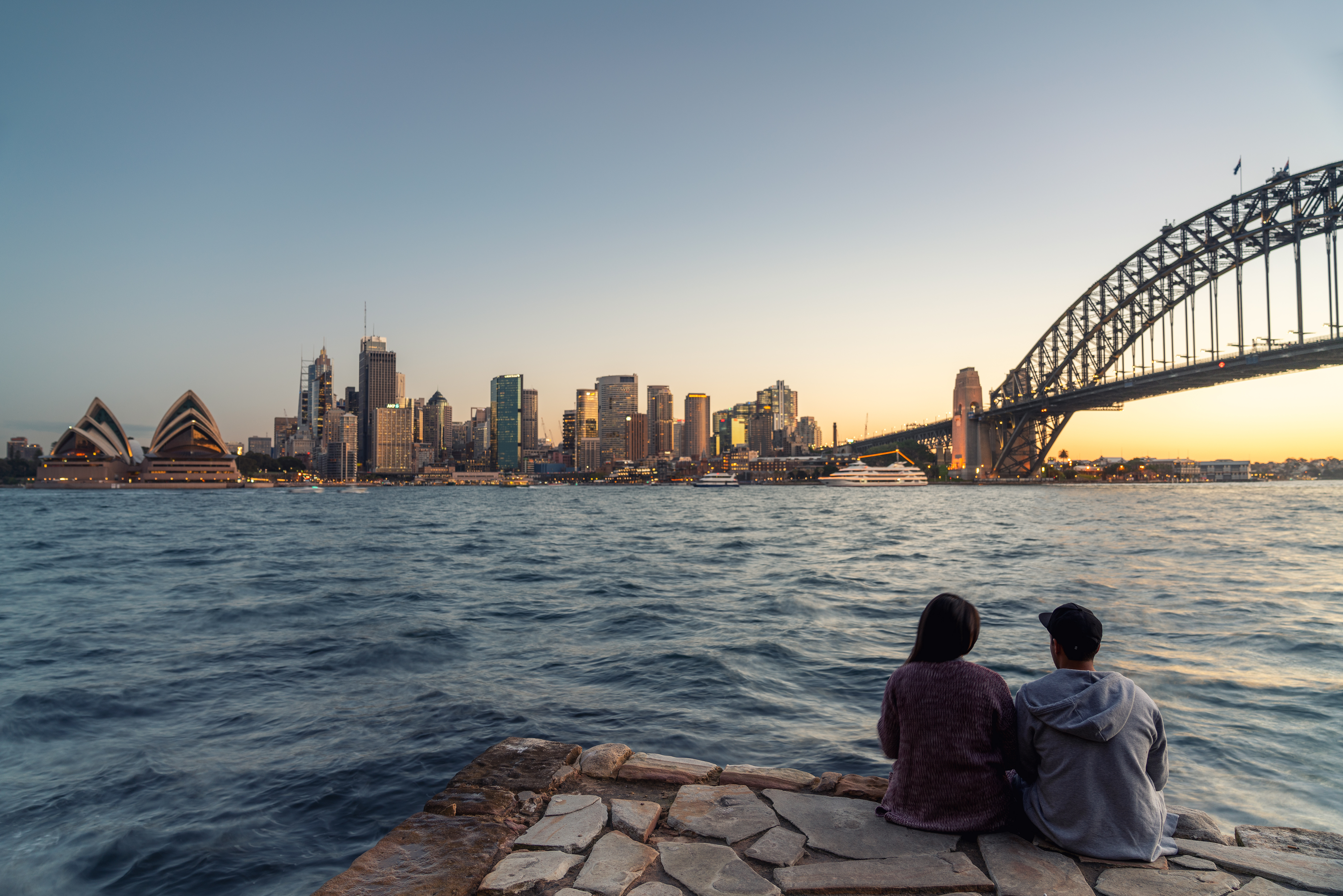 couple sitting on edge of harbour looking at bridge and skyline