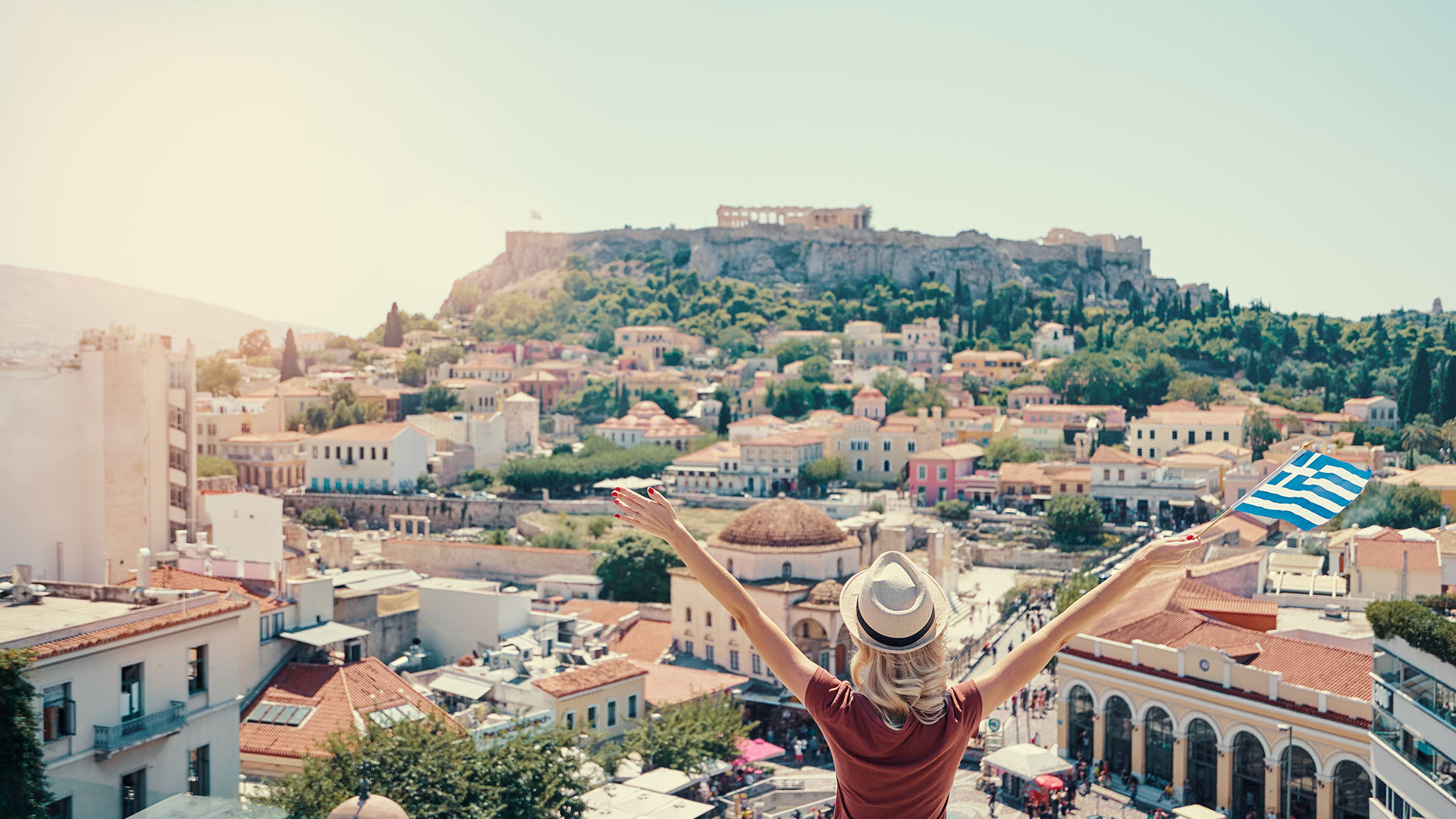 A woman stretching her arms out with a Greece flag in hand, looking out across Athens to Acropolis on the horizon