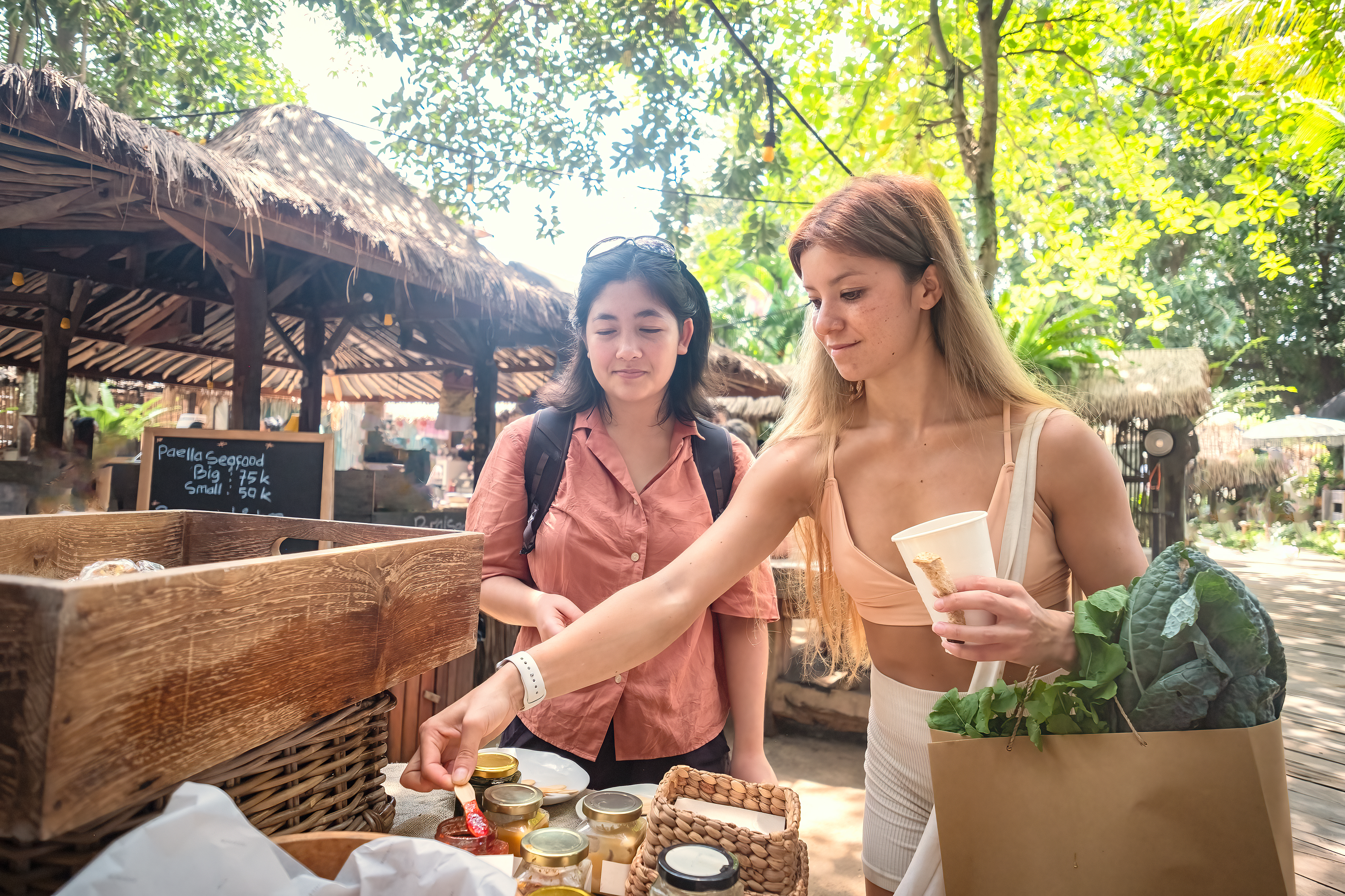 Multiracial young woman nutritionist shopping with Asian client for organic vegan food at a public outdoor market in Canggu, Bali, Indonesia. Nutritionist is holding paper shopping bag containing arugula and kale.