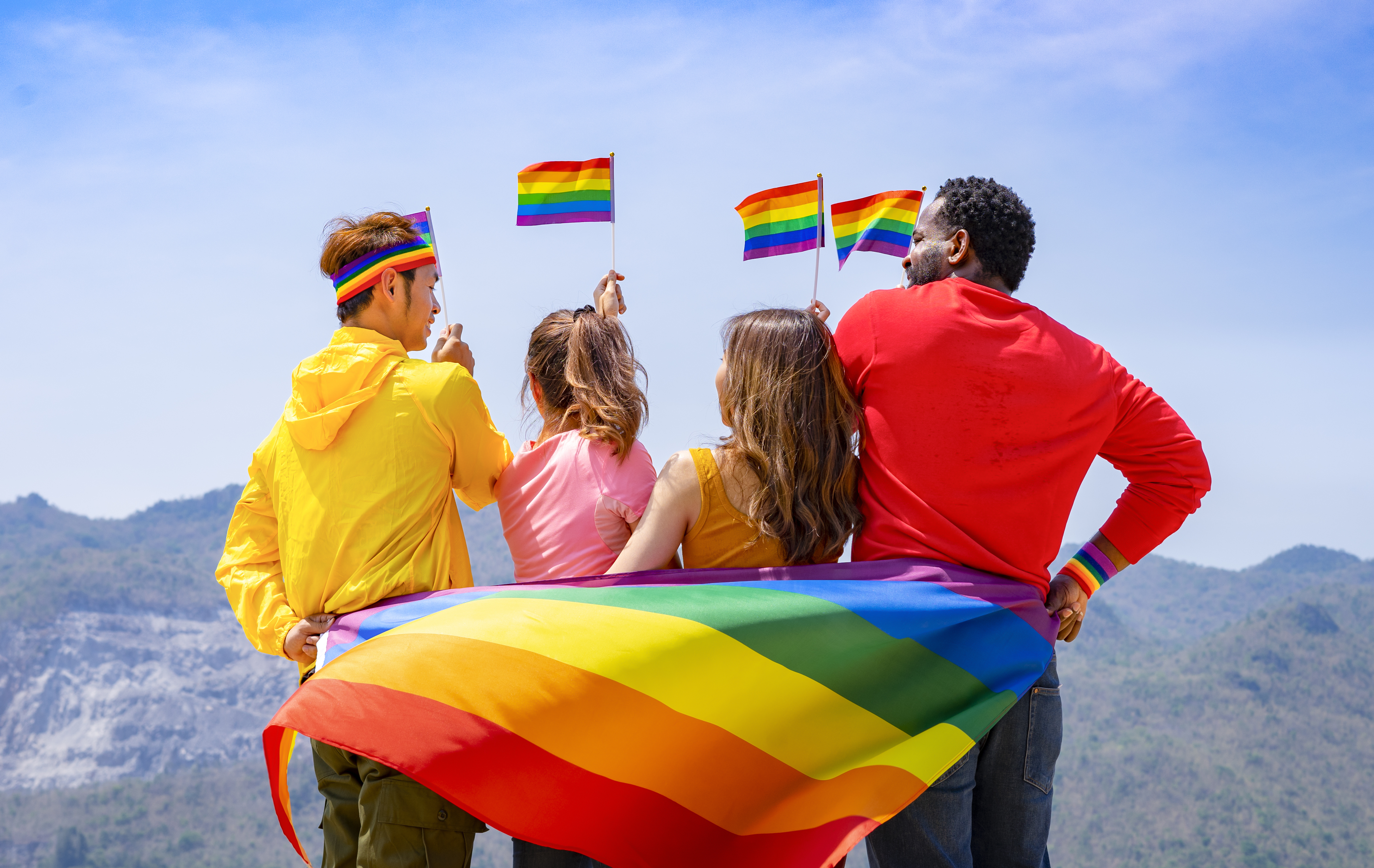 Group of diverse lgbtq friends wearing colorful clothes, standing side by side, smiling and holding lgbt flags waving in the air