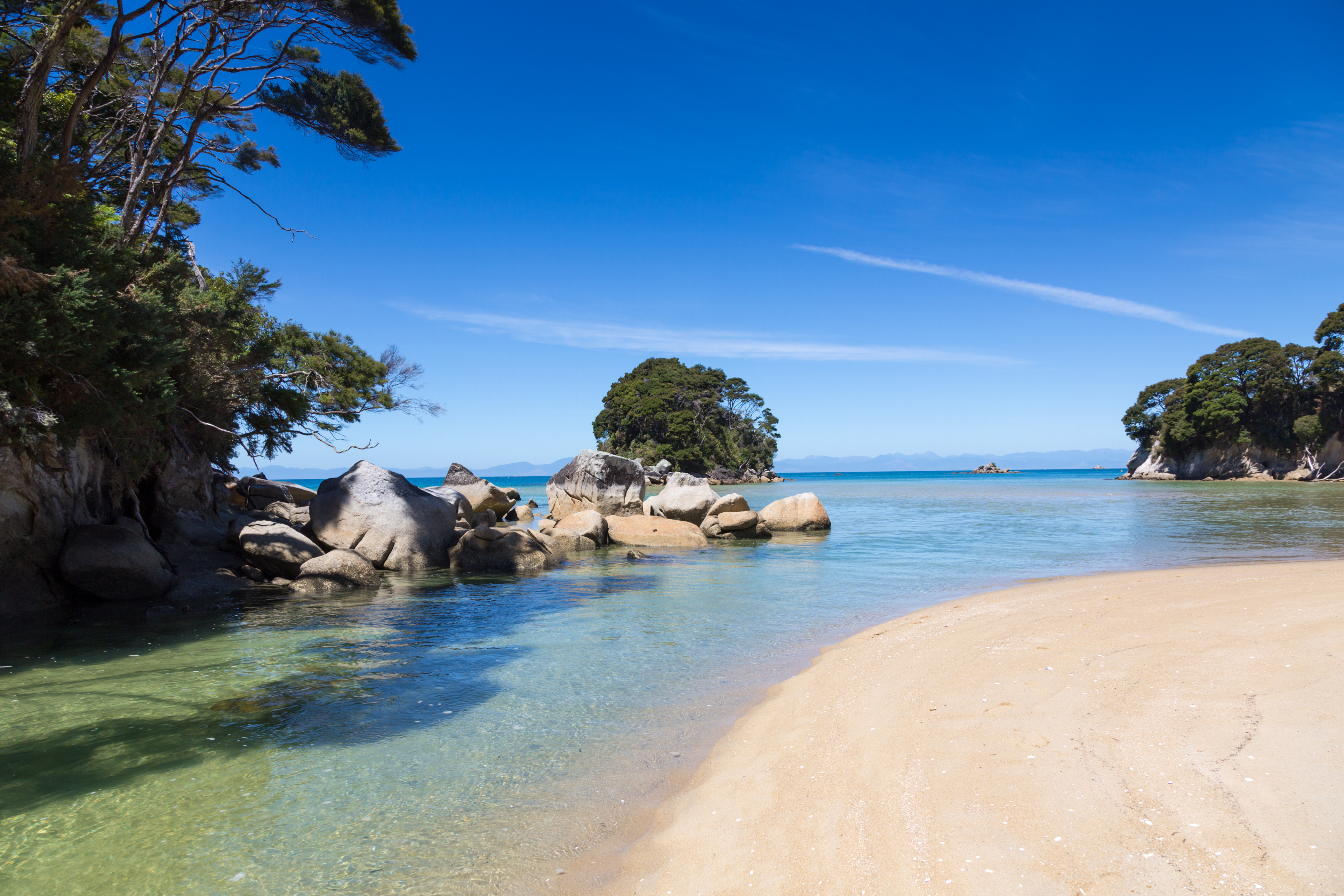 Abel Tasman National Park on South Island New Zealand on the beach on day time. The water is calm and clear. There is big rocks into the water and few islands. The sky is clear blue.