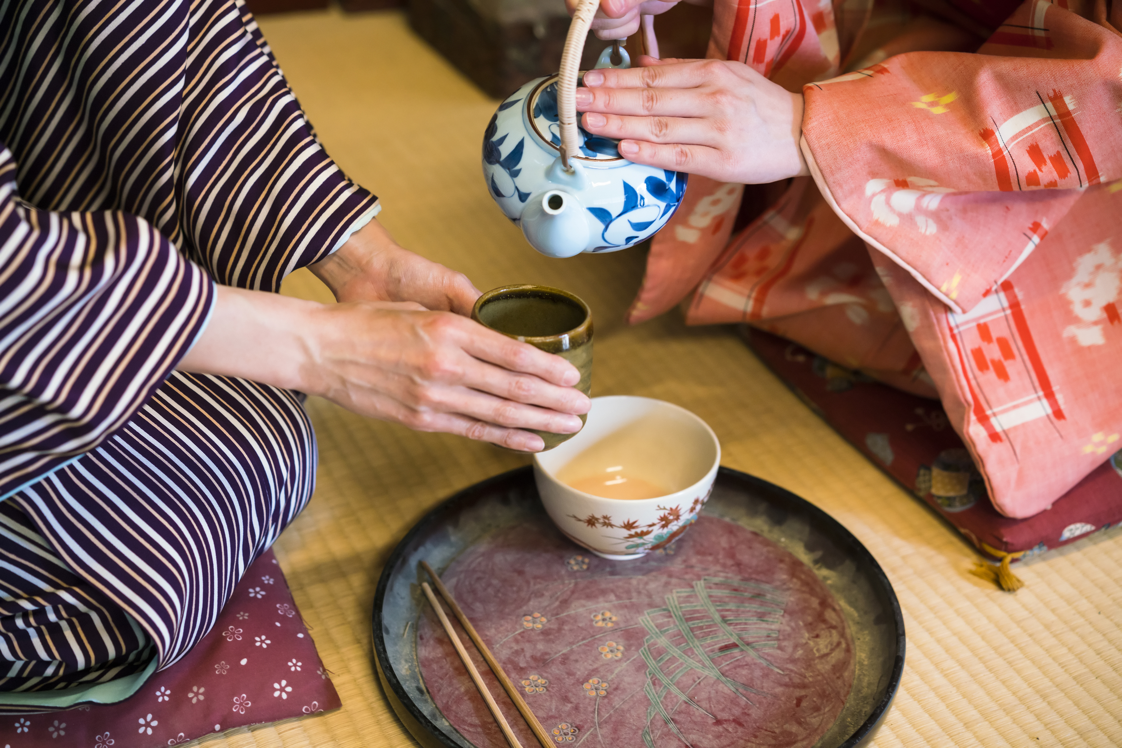 detail of two women in traditional kimono, kneeling on tatami preparing, pouring a cup of tea which is inthe hands of one woman. They are in traditional Japanese old house on tatami. This is in Toei studios in Kyoto with old buildings from Samurai times.
