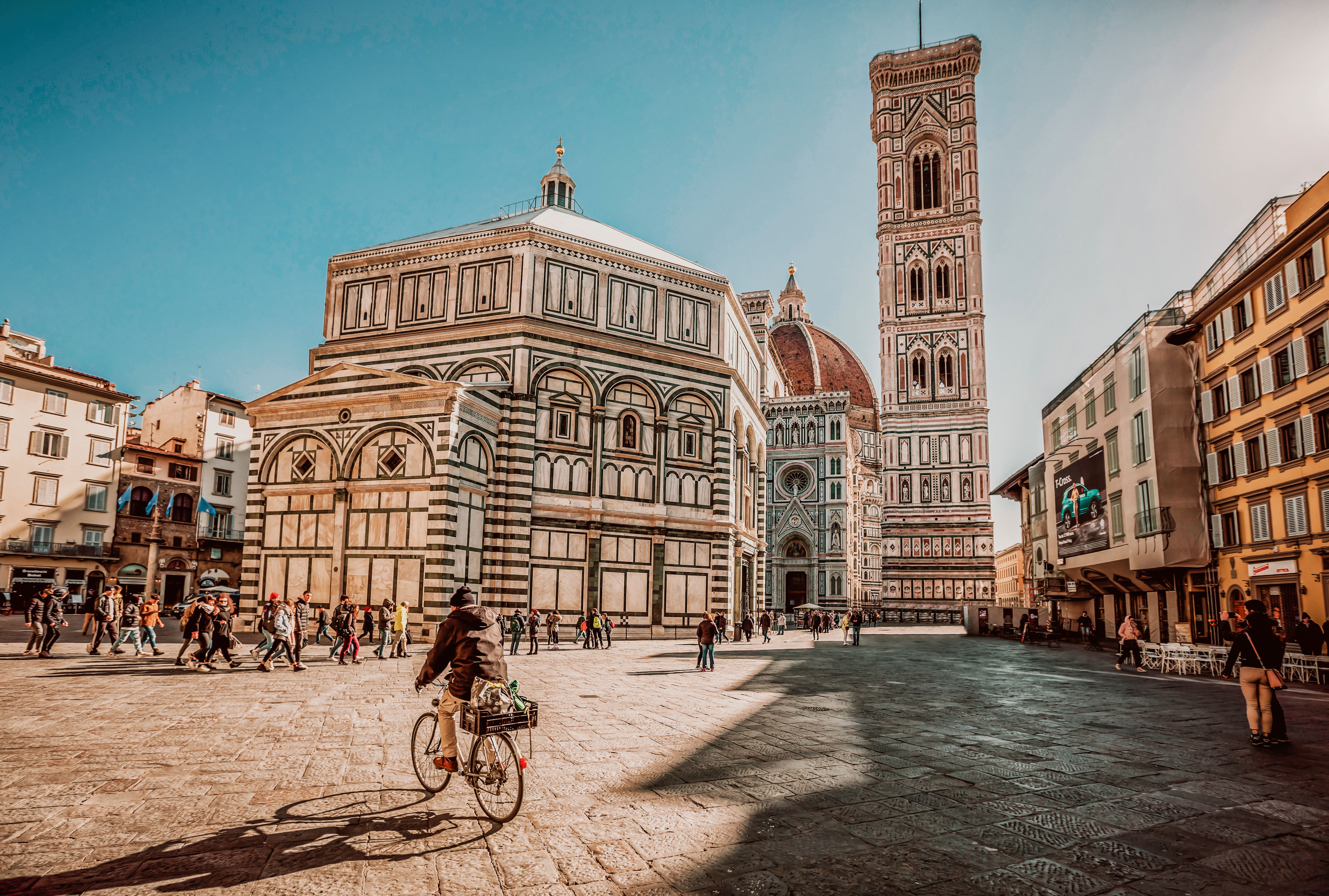 a man rides a bike into the charming old square of Piaza del Duomo in Florence, where the massive cathedral looms in the background with an ever-present crowd.