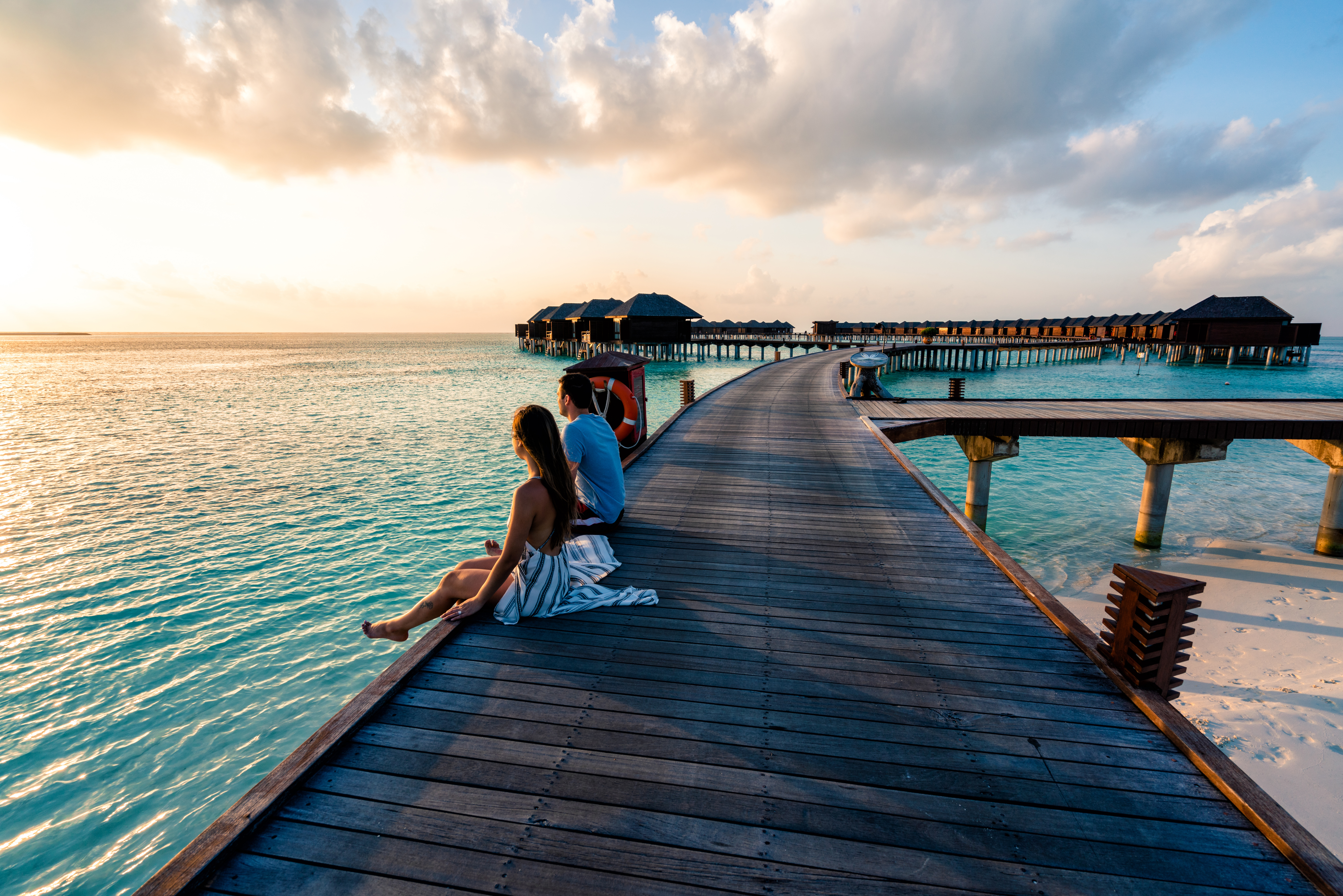 A couple sitting on a boardwalk leading out into the water in the Maldives.