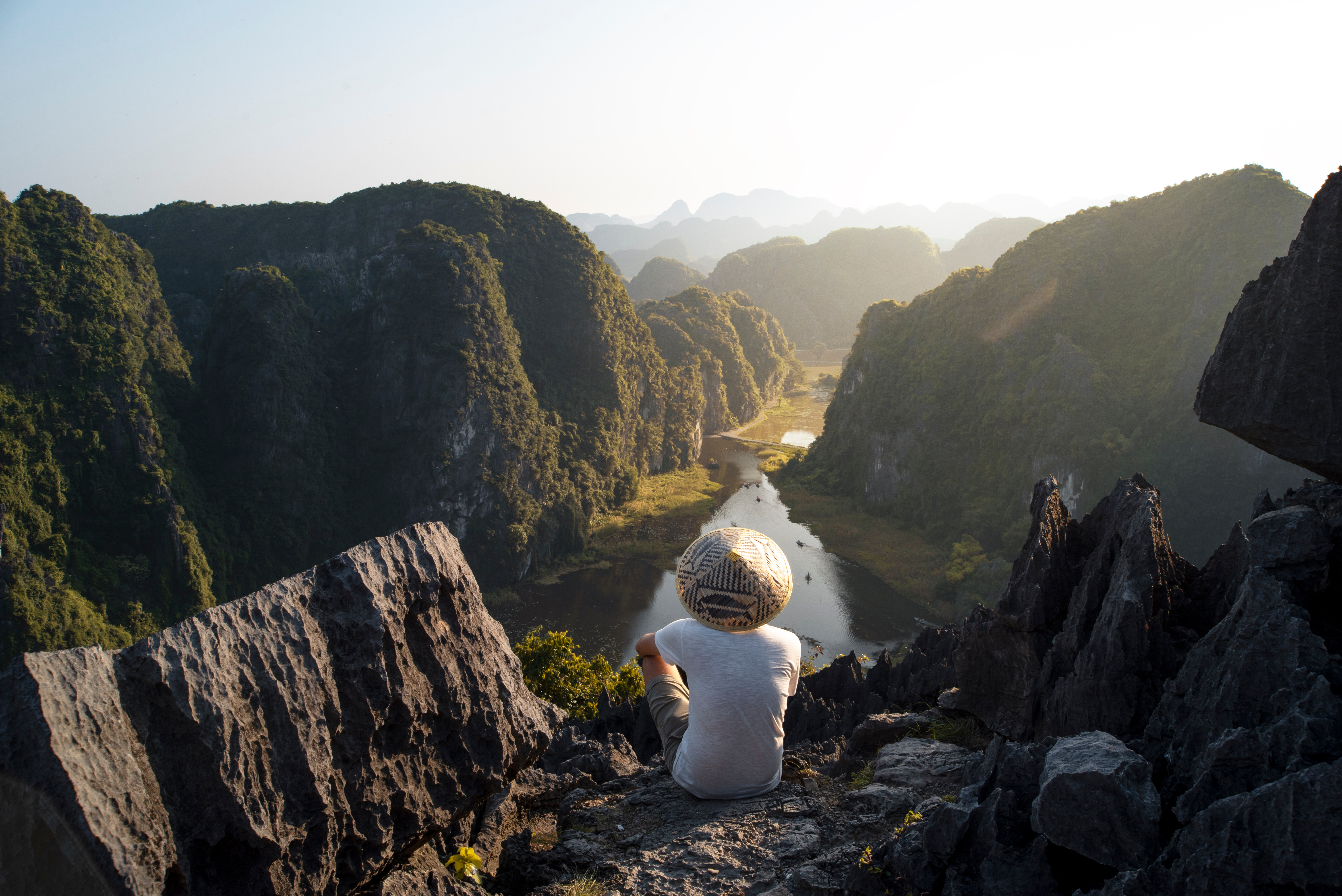 man in a straw hat sitting on the edge of a mountain overlooking river