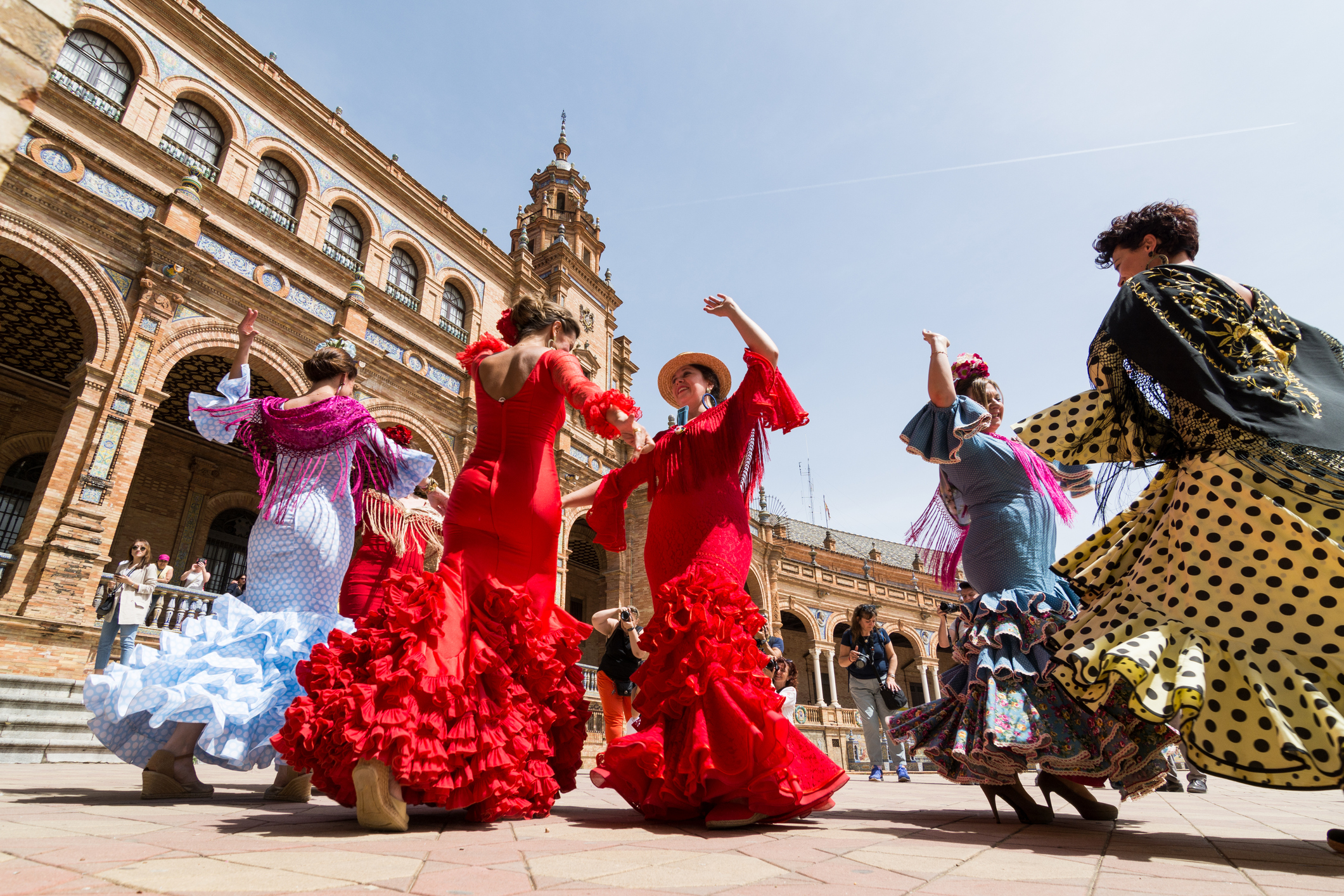 A group of women in colorful flamenco dresses dance in a plaza with historic architecture in the background.