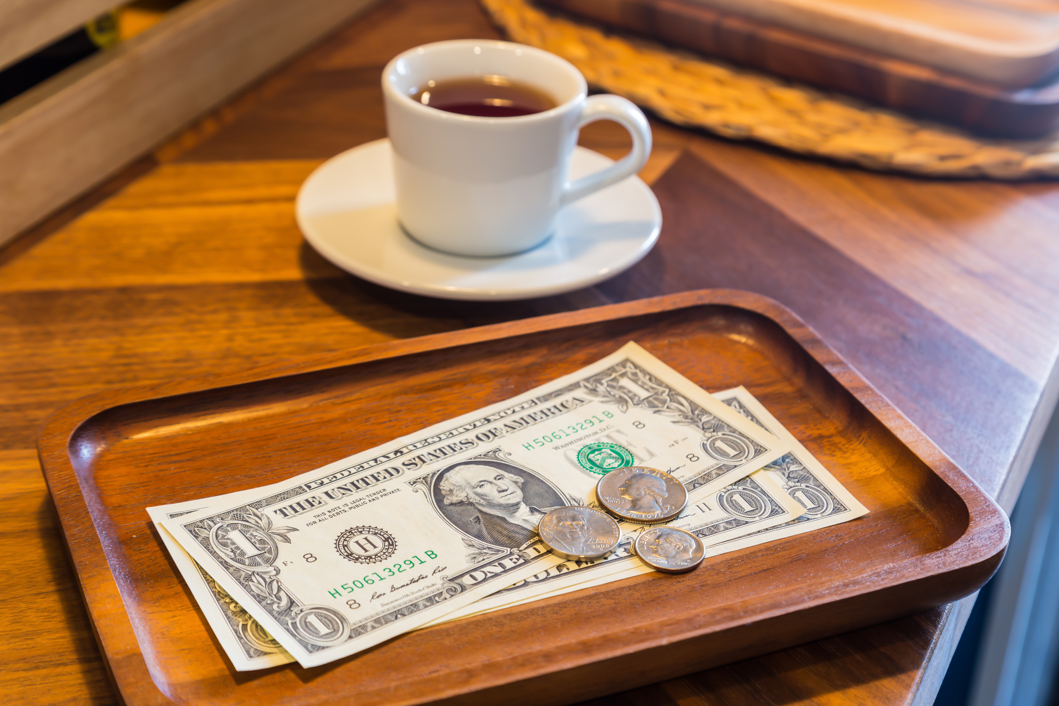 A wooden tray holds a dollar bill and coins, beside a white cup of tea on a wooden table.