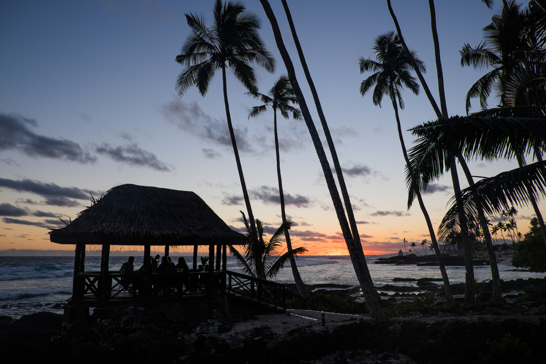 Silhouette of a beach pavilion at sunset, surrounded by palm trees and ocean waves.
