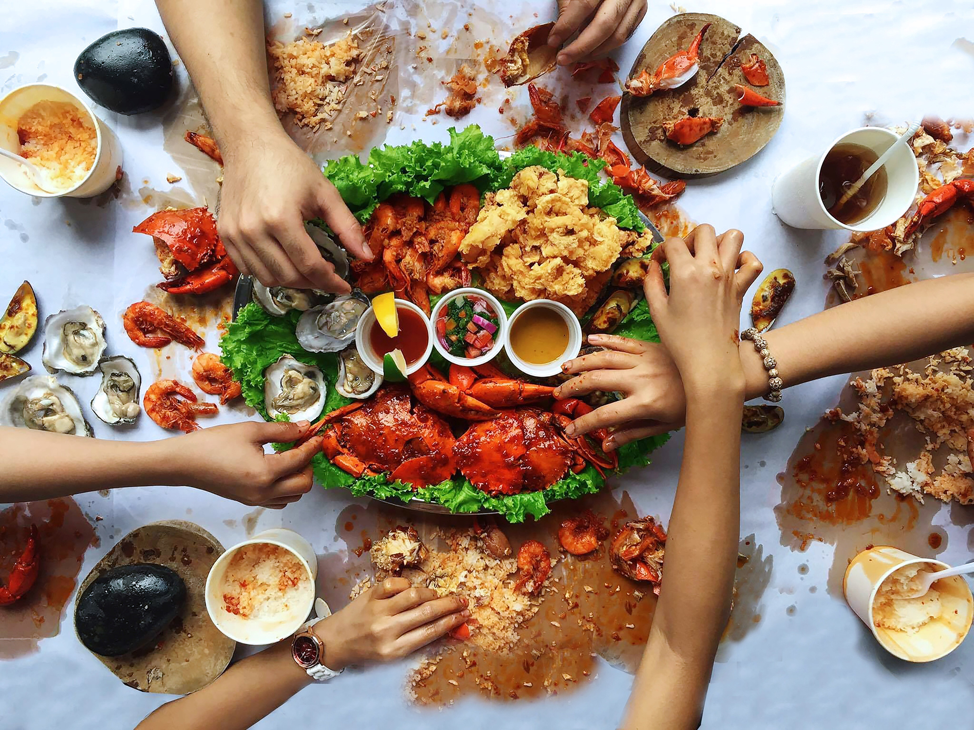A feast of seafood, including lobsters and oysters, with hands reaching for food on a messy table covered in shells and sauces.