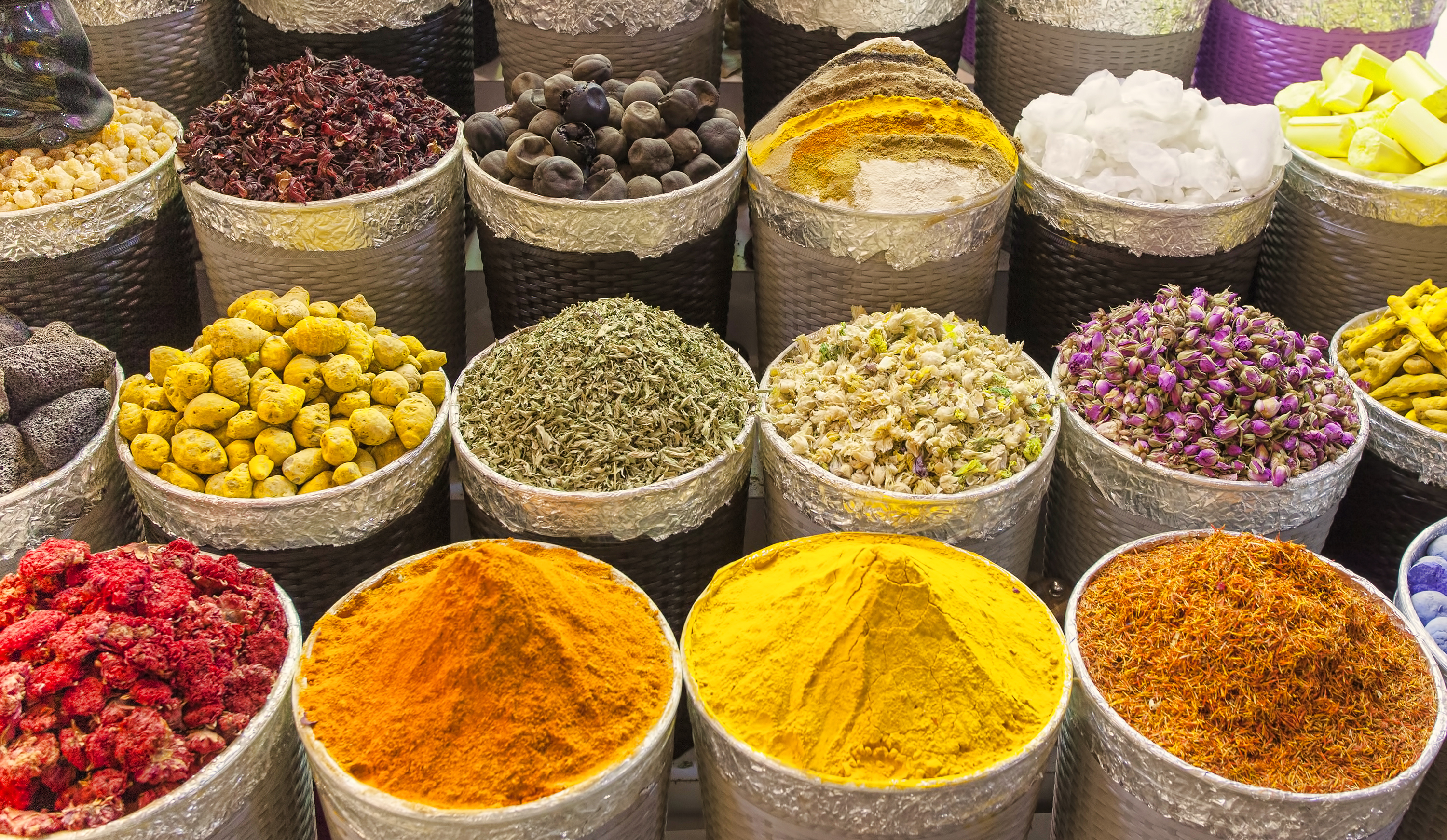 Colorful display of spices in metal containers, including saffron, turmeric, dried herbs, and floral petals.