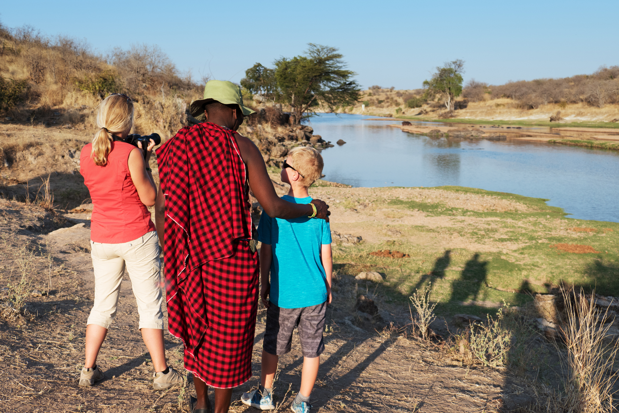 A woman and a boy watch a river with a man in traditional attire, capturing the moment as the sun sets.
