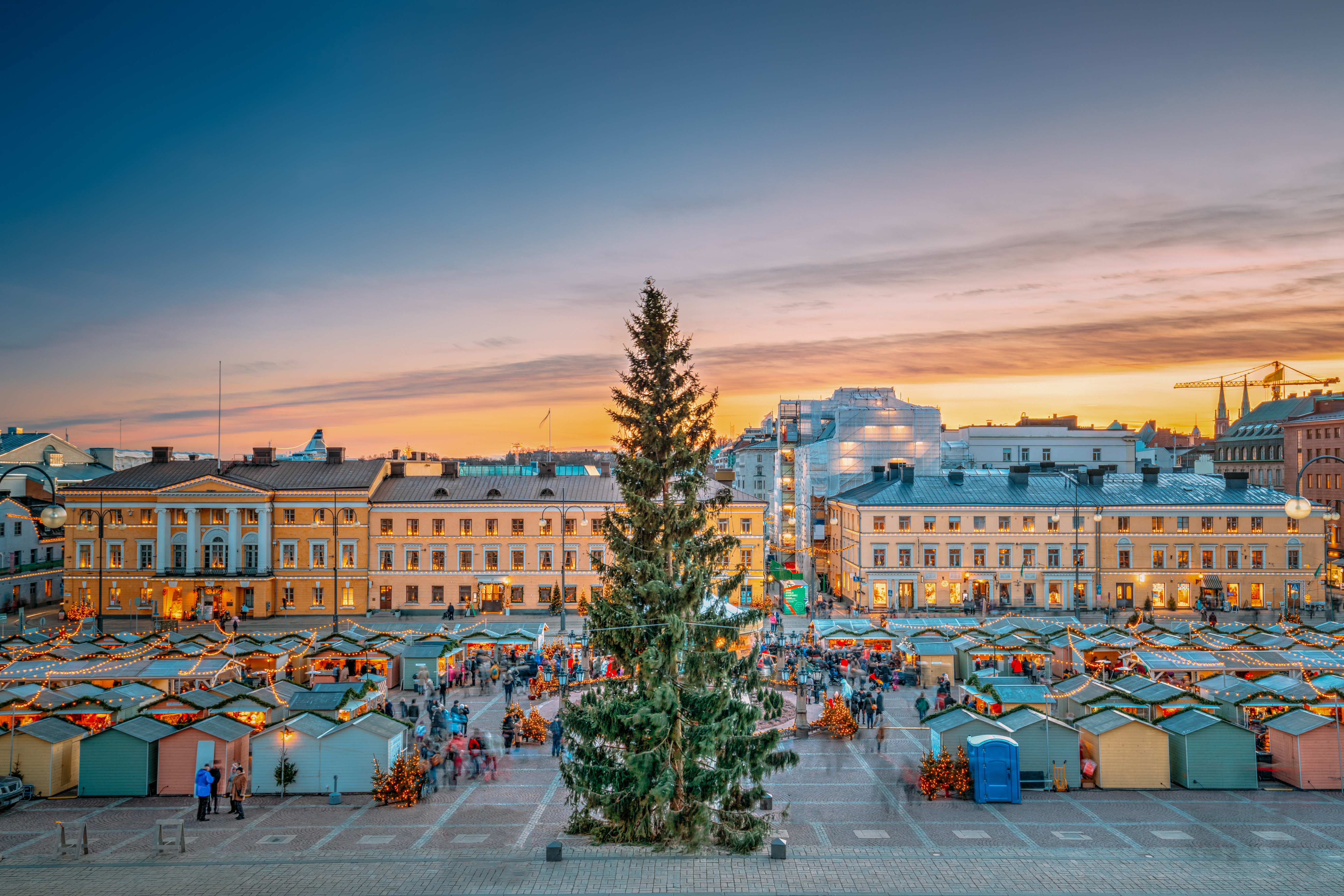 A vibrant market square at sunset, featuring a large Christmas tree and festive stalls filled with visitors and decorations.