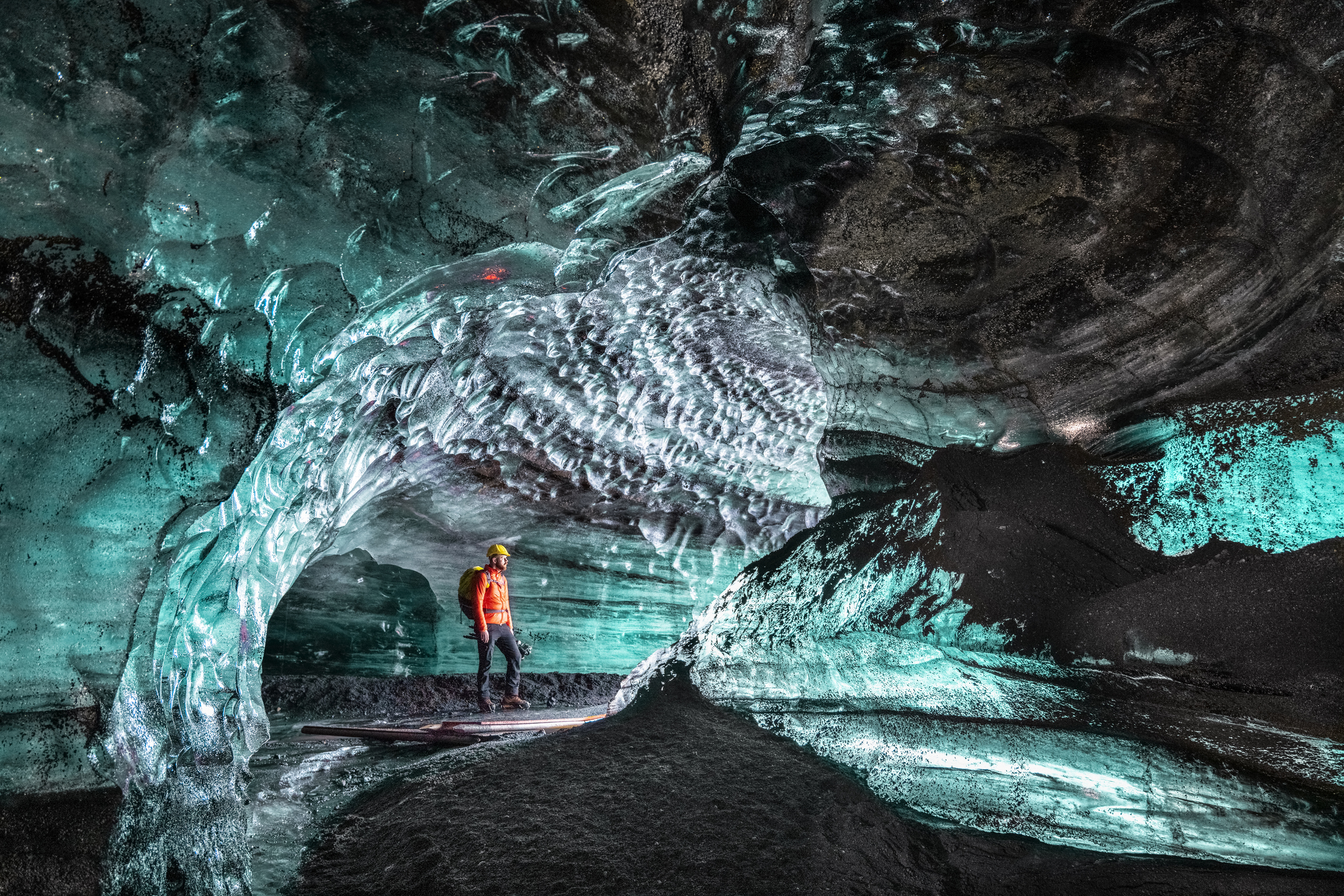 A person in an orange jacket stands inside a stunning ice cave with textured blue ice formations and intricate patterns.