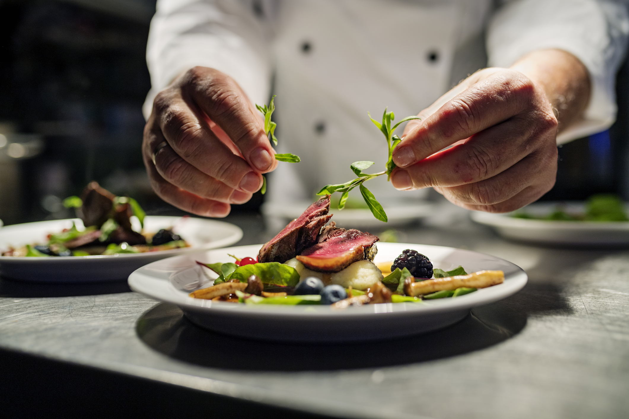 Chef garnishing a beautifully plated dish with fresh herbs in a restaurant kitchen.