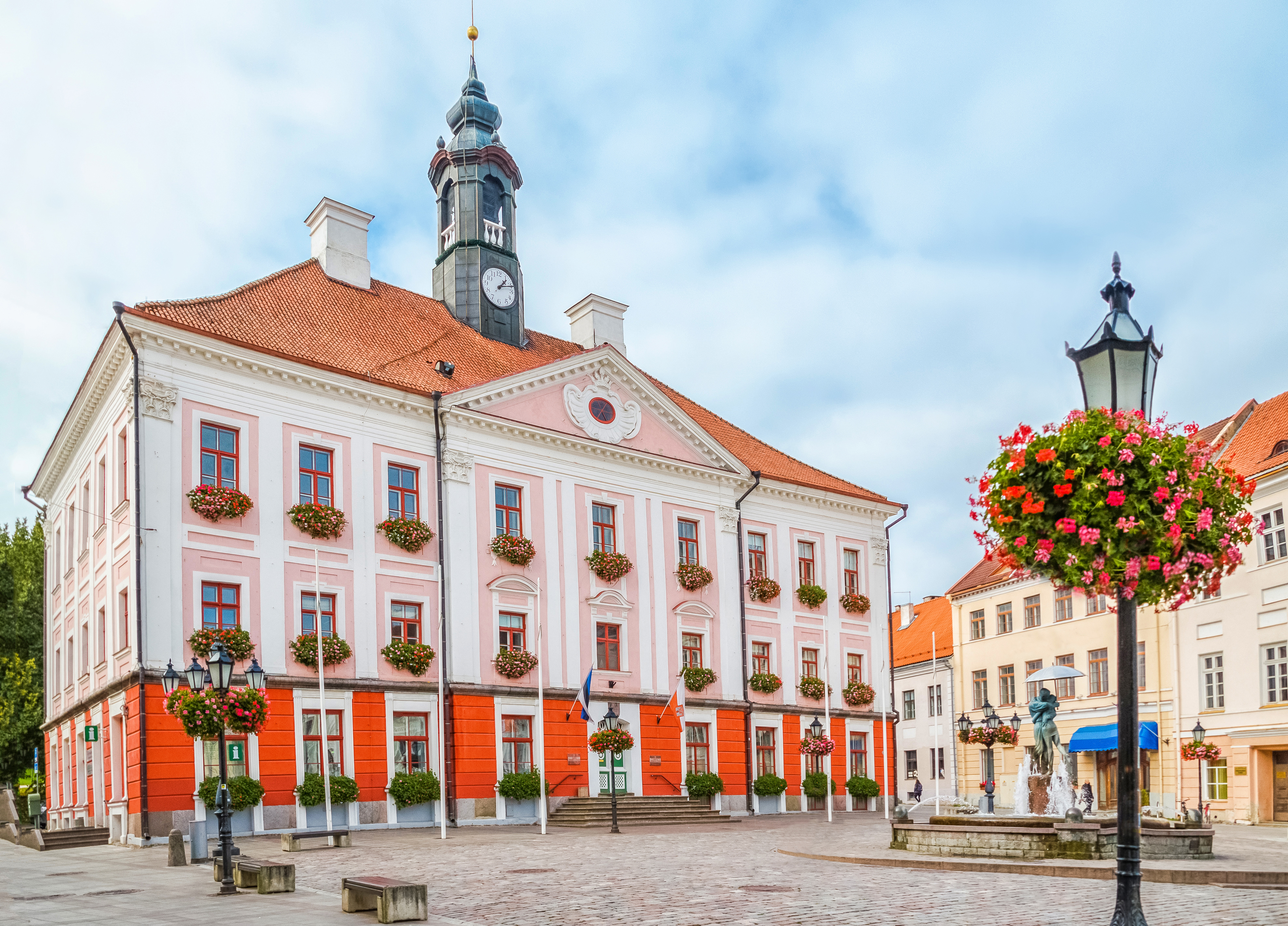 Pink and white town hall with a tower, surrounded by blooming planters and a cobblestone square on a cloudy day.