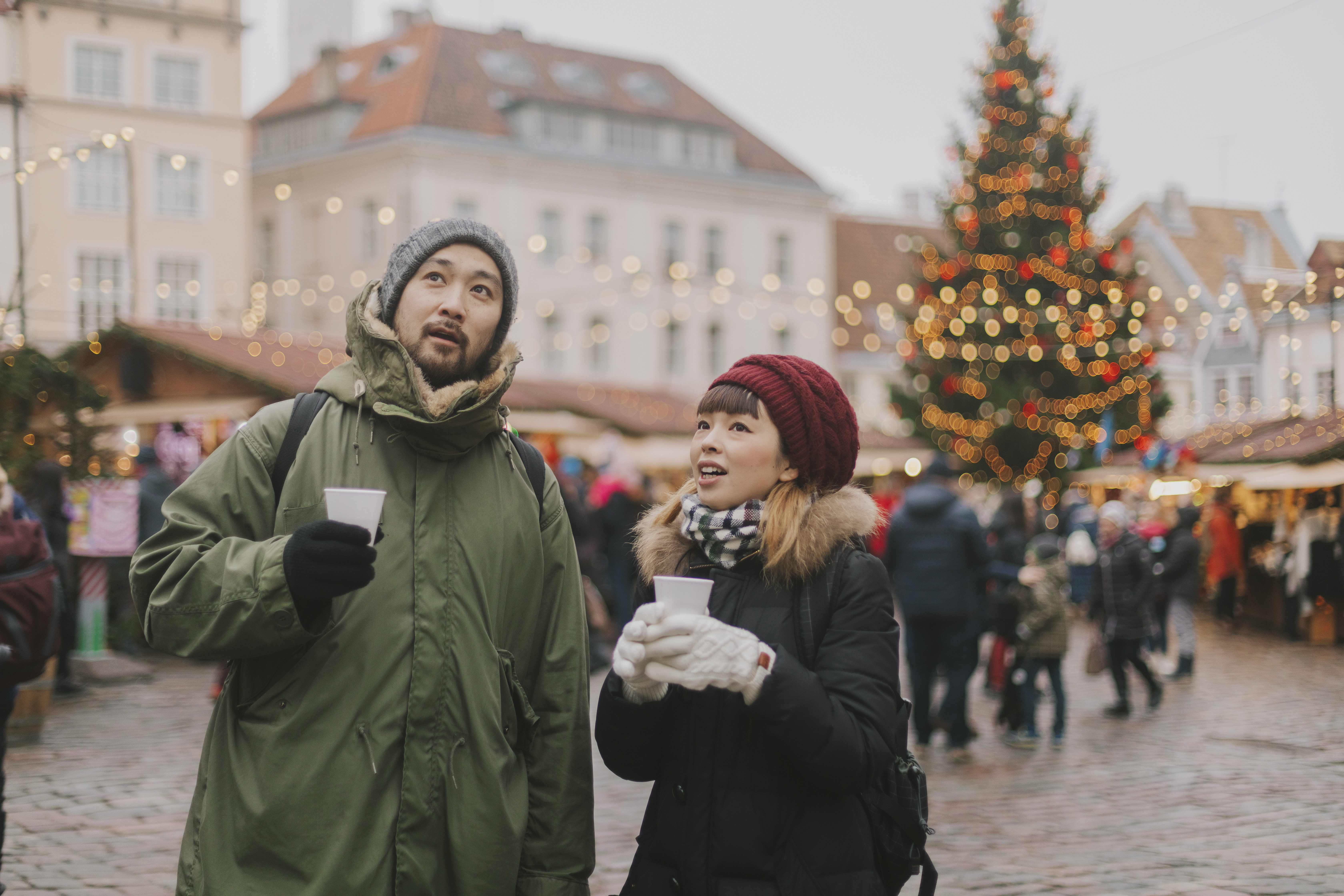 A man and woman look up, holding cups, in a festive market with holiday lights and a Christmas tree in the background.