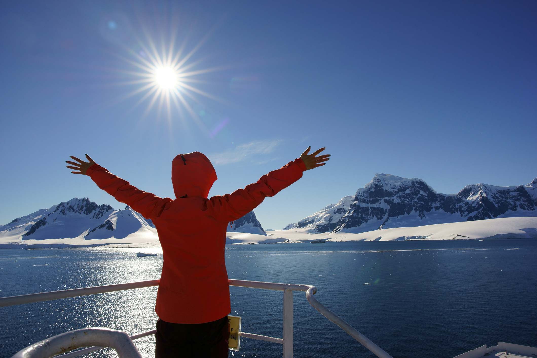 Person in a red jacket spreading arms wide, facing sunlit mountains and ocean under a clear blue sky.