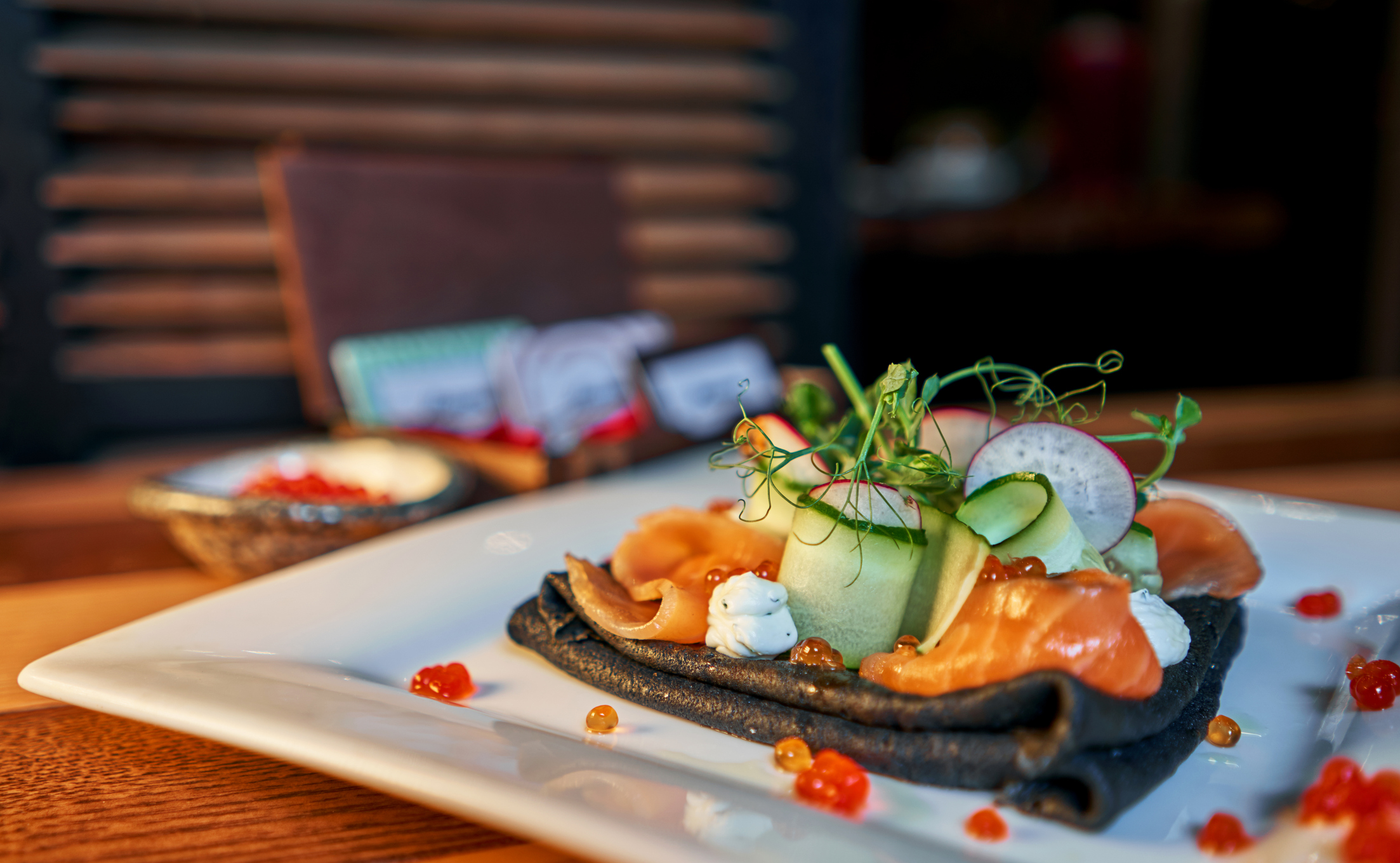 A gourmet dish with black crepes, smoked salmon, cucumber, cream, and orange caviar, elegantly plated on a white dish.
