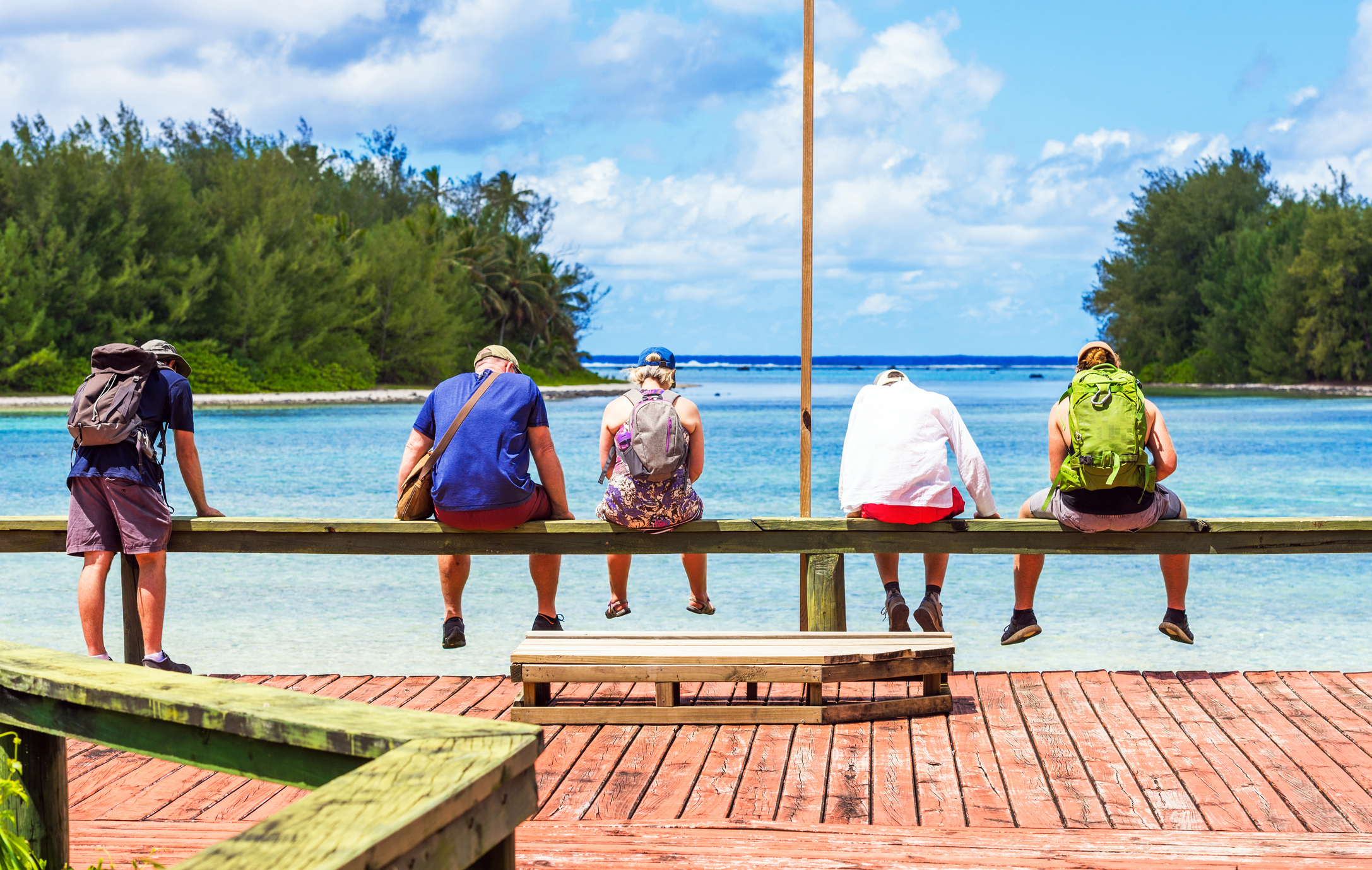 Five people sit on a wooden railing by the water, facing the ocean under a blue sky with scattered clouds.