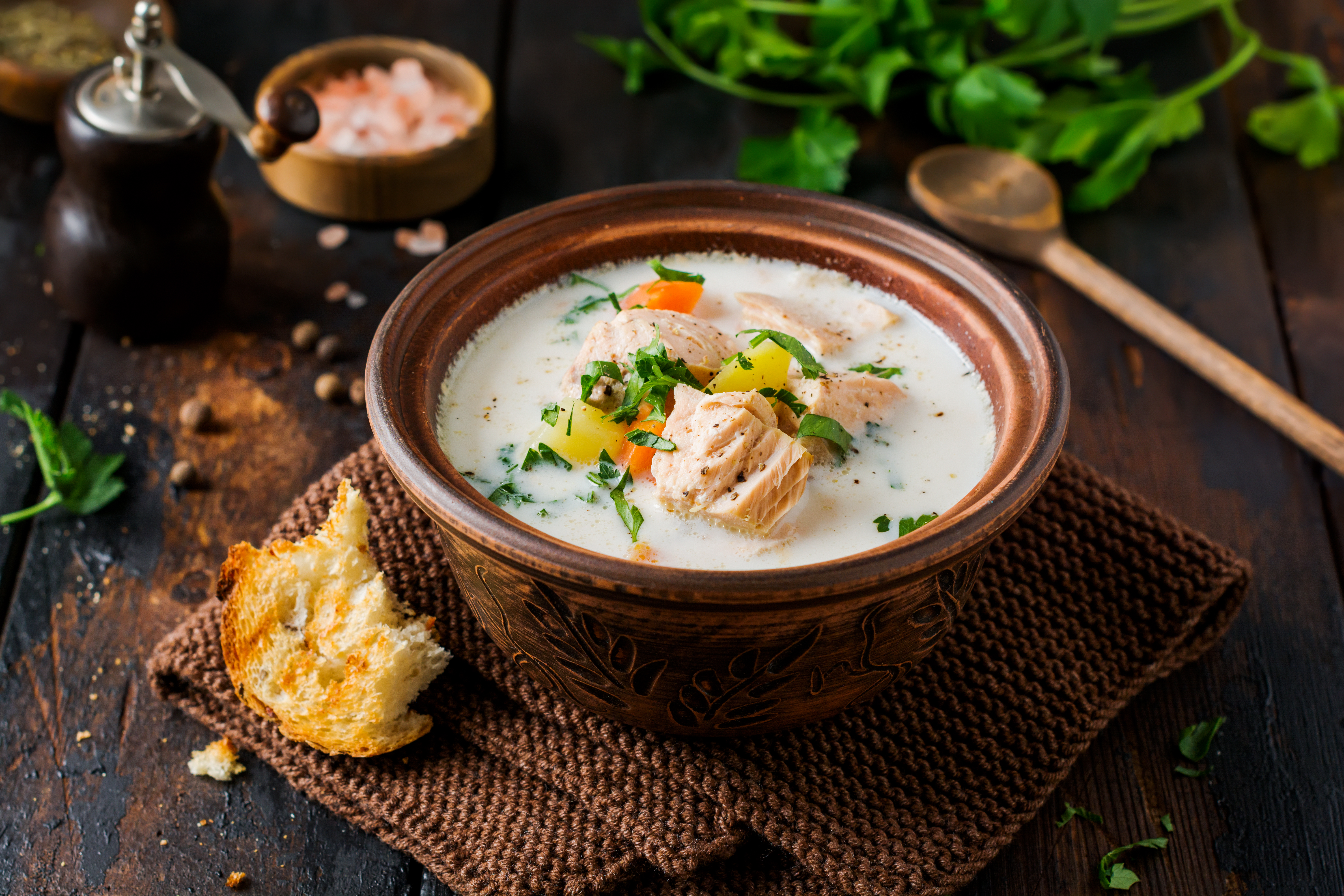 Bowl of creamy salmon soup with vegetables, garnished with herbs, beside a piece of bread on a woven mat.