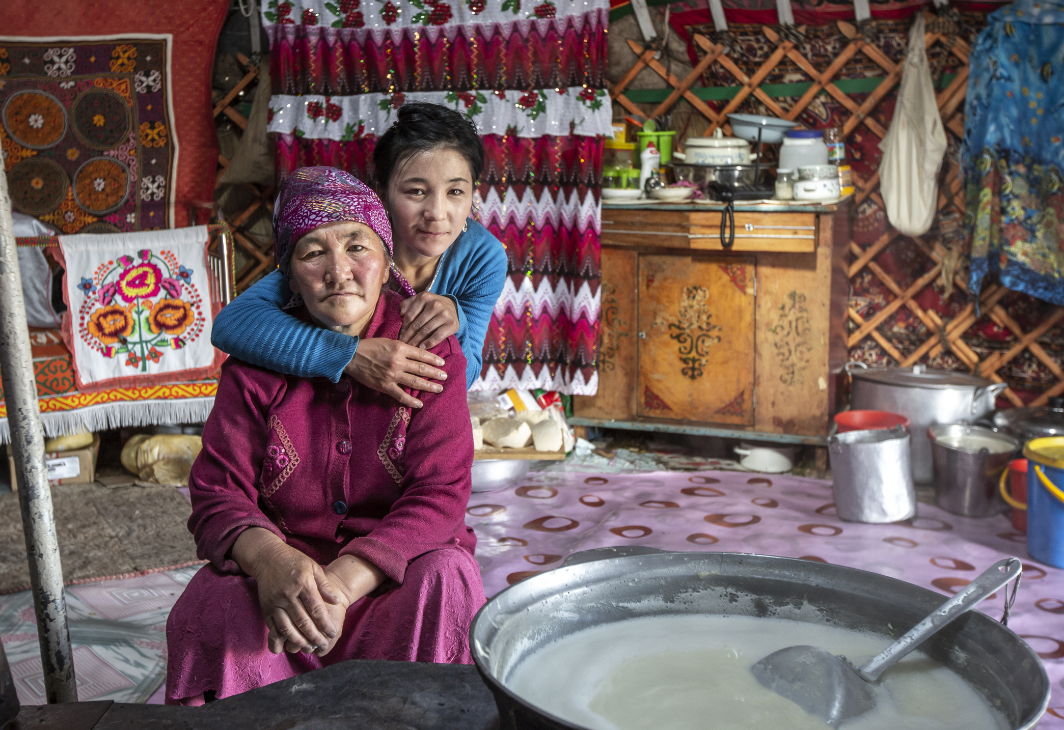 A young girl embraces an elderly woman in a cozy, traditional yurt adorned with colorful textiles and kitchen items.