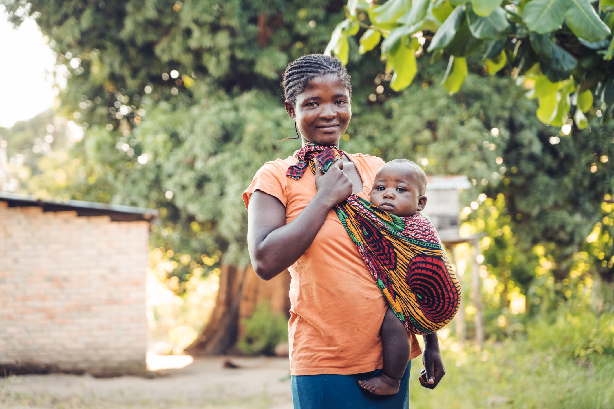 A woman wearing an orange top holds a baby wrapped in colorful fabric, standing outdoors near a home with trees in the background.