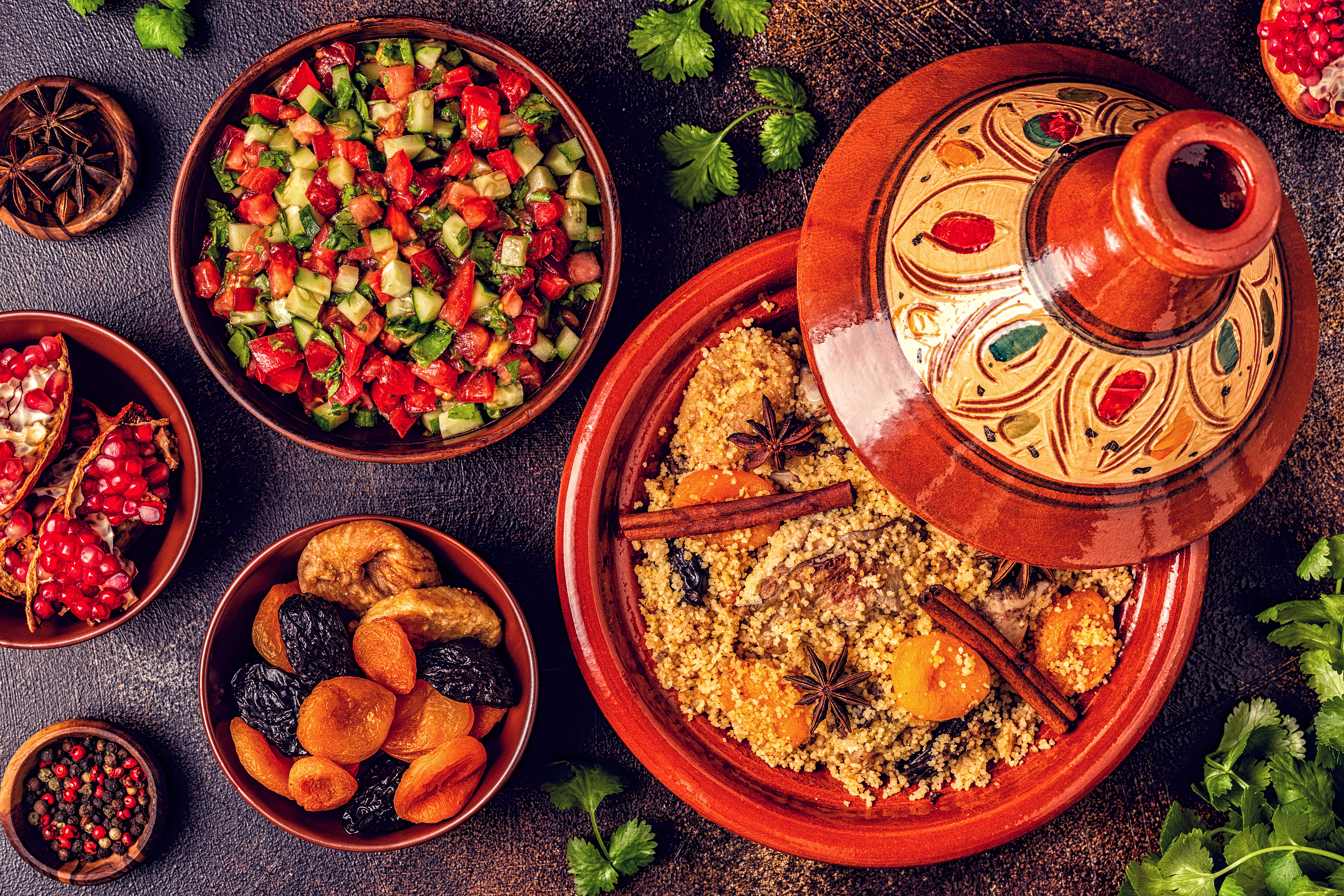 A colorful spread of couscous with spices, surrounded by salads, dried fruits, and garnishes on a textured surface.