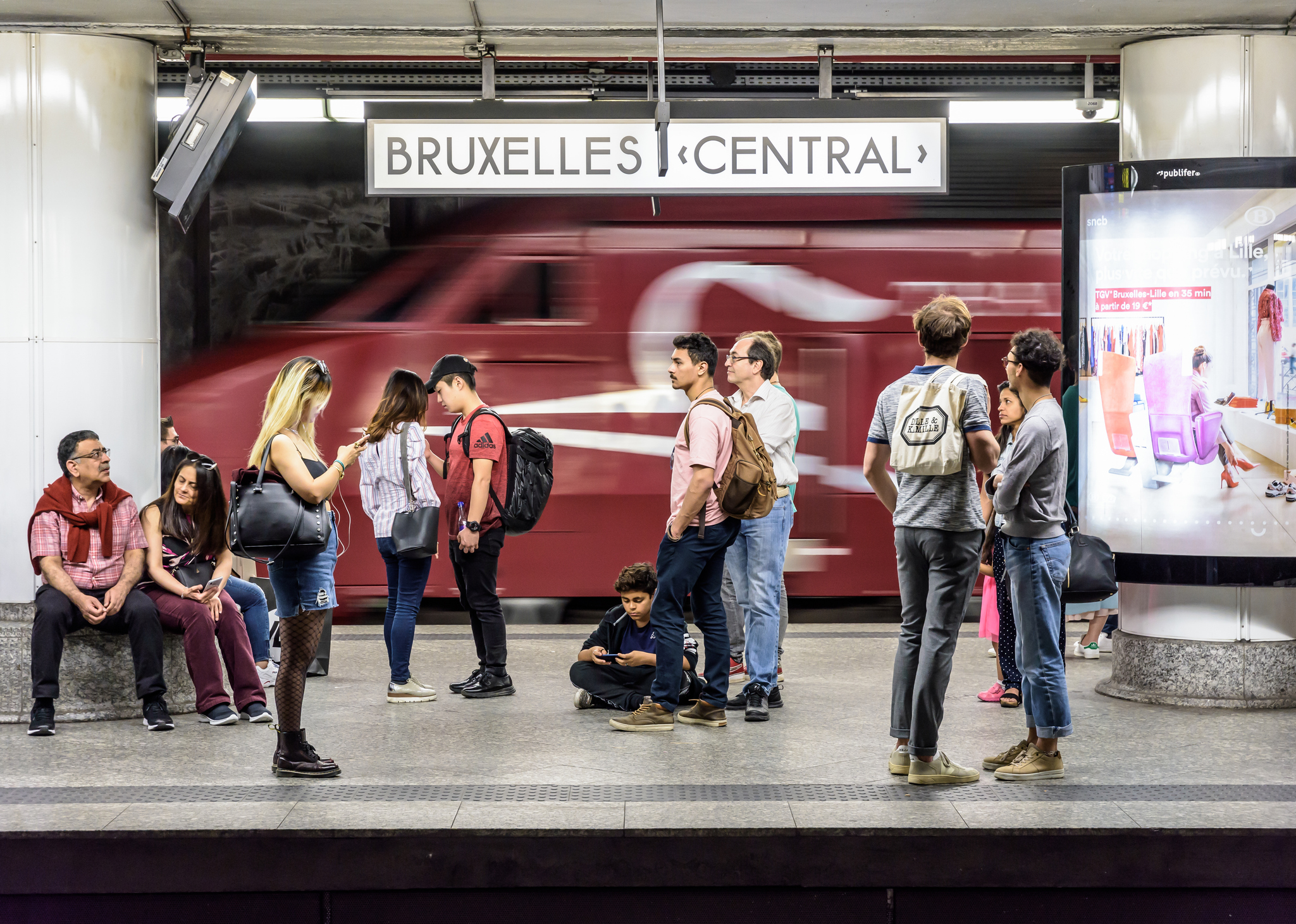 A busy train station platform at Bruxelles Central, with passengers waiting and a train passing in the background.