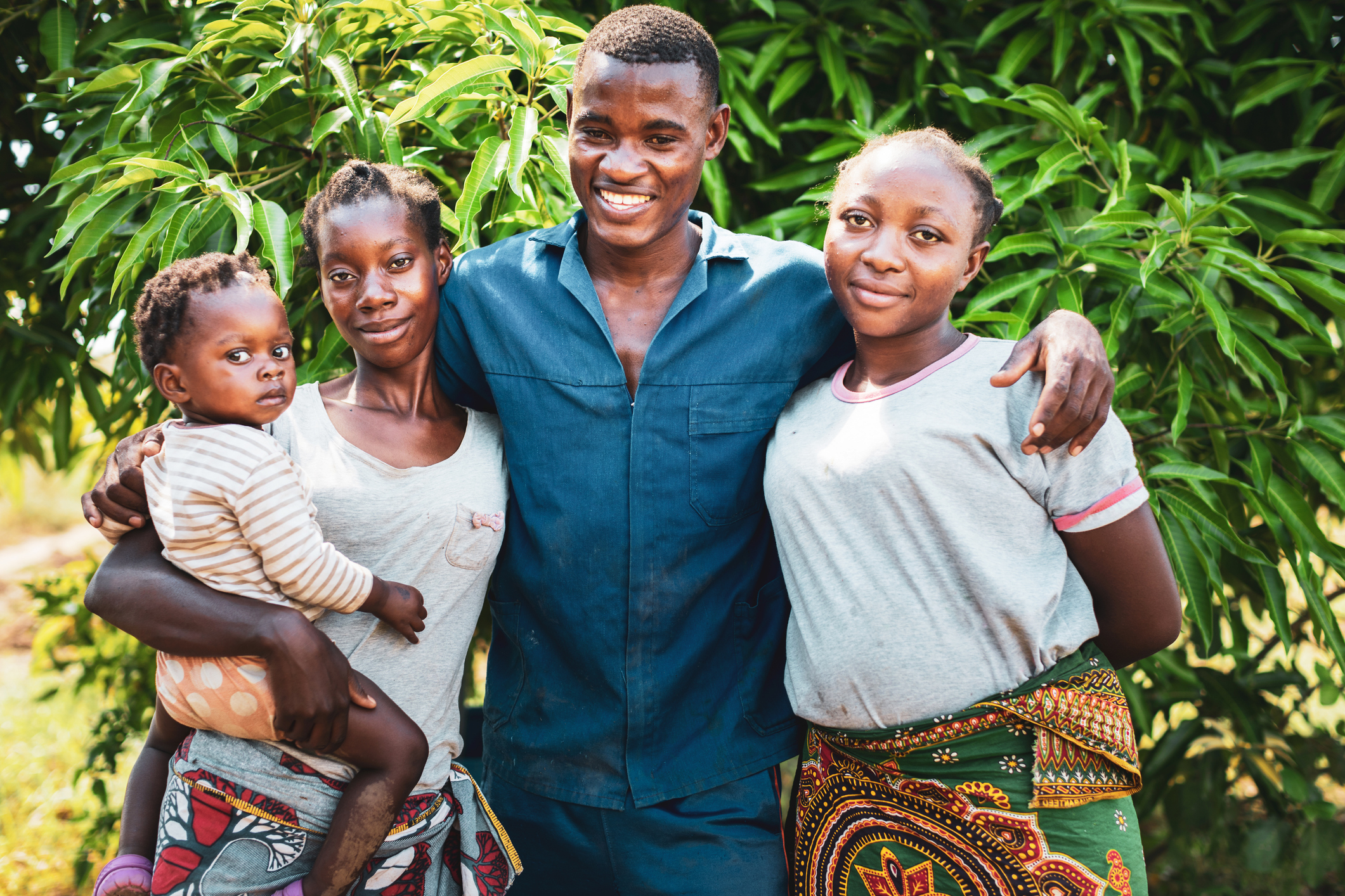 A smiling man stands with three women, one holding a baby, in front of lush green foliage.