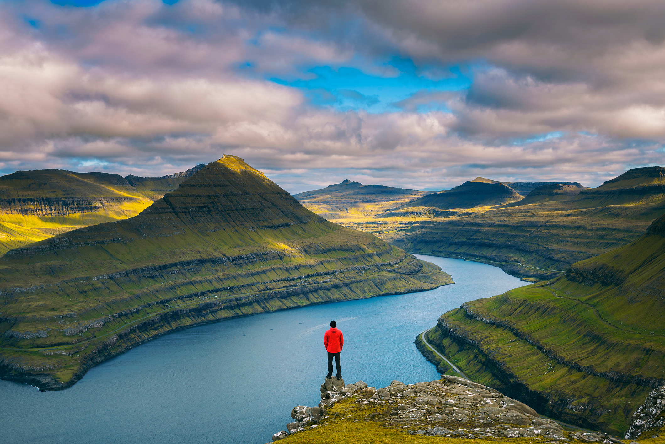 A person in a red jacket stands on a cliff overlooking a winding river and lush green mountains under a cloudy sky.