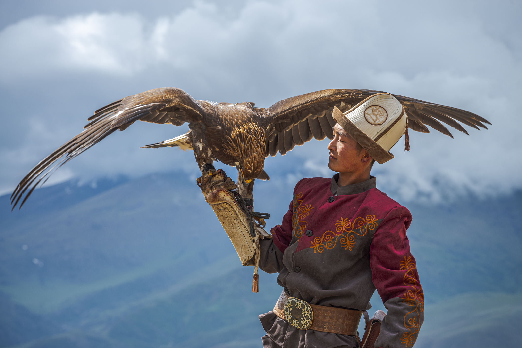 A man in traditional attire holds a large eagle on his arm against a mountainous backdrop.