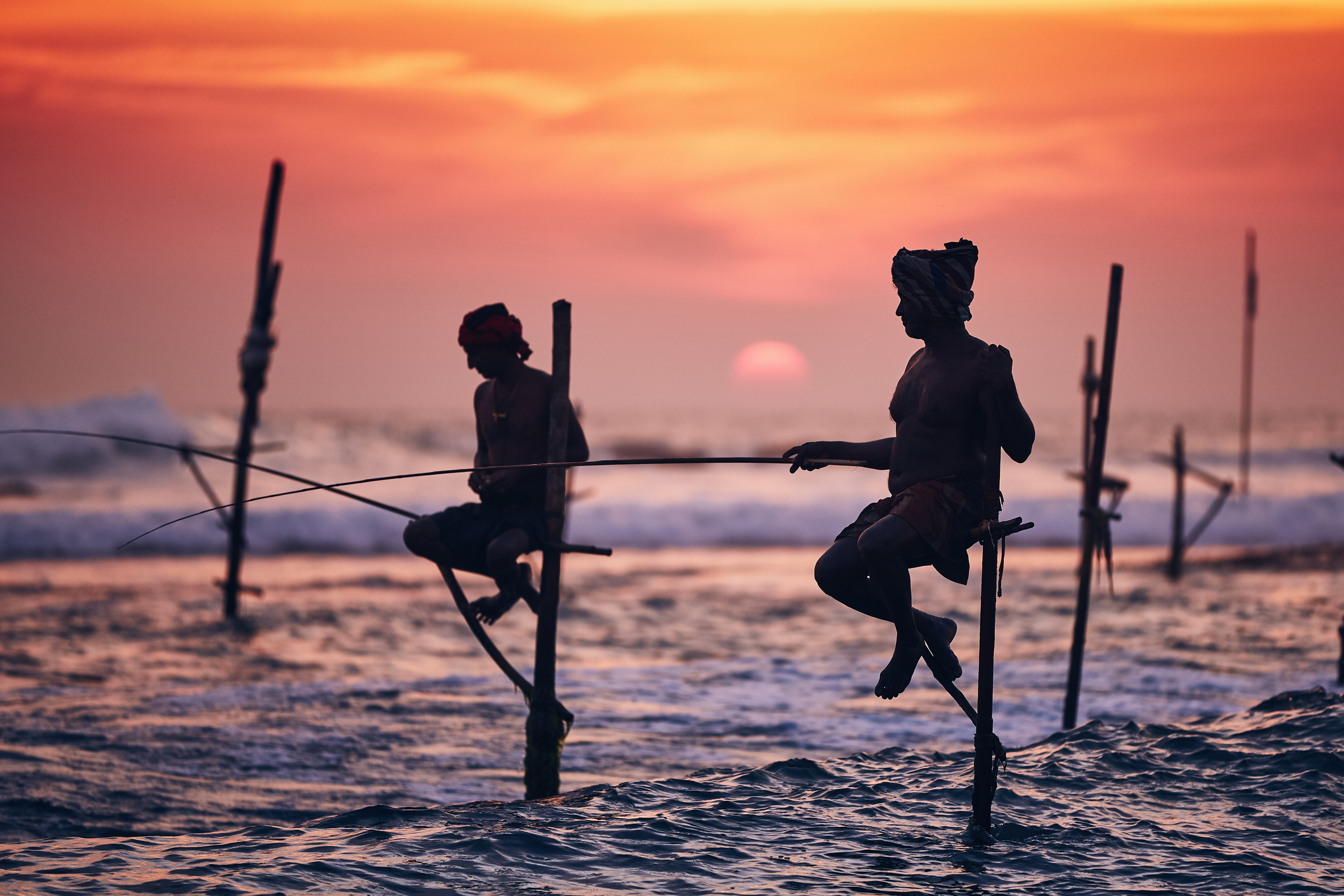 Silhouetted fishermen perched on poles in the ocean at sunset, casting lines into the water.