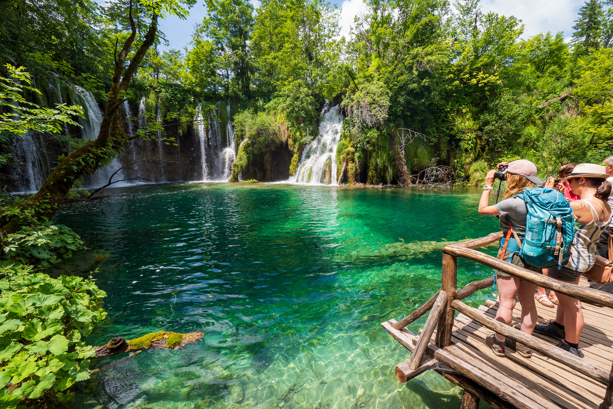 Two people stand on a wooden platform, taking photos of a turquoise lake and waterfalls surrounded by lush greenery.