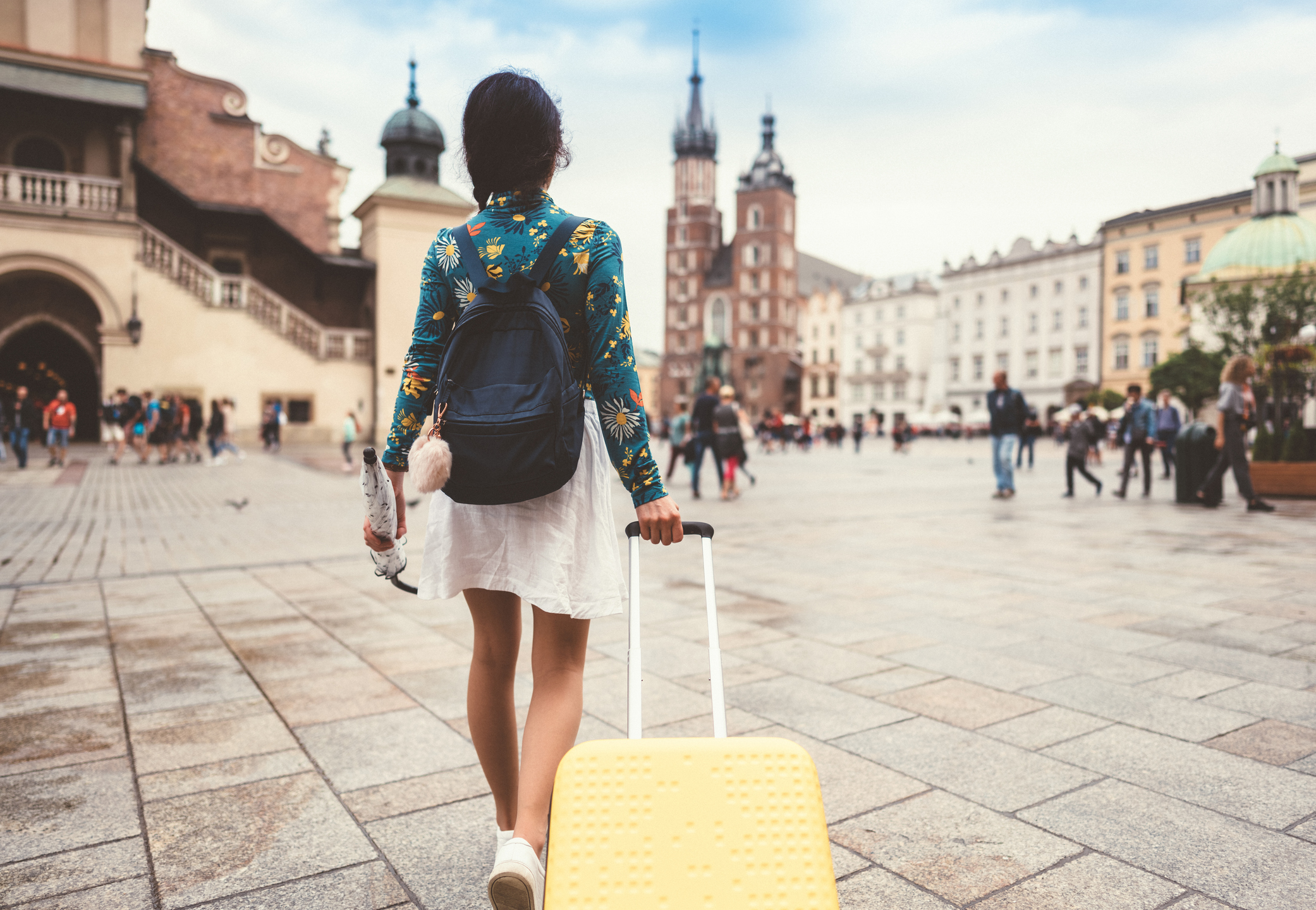 A person walks away, pulling a yellow suitcase through a bustling square with historic buildings in the background.
