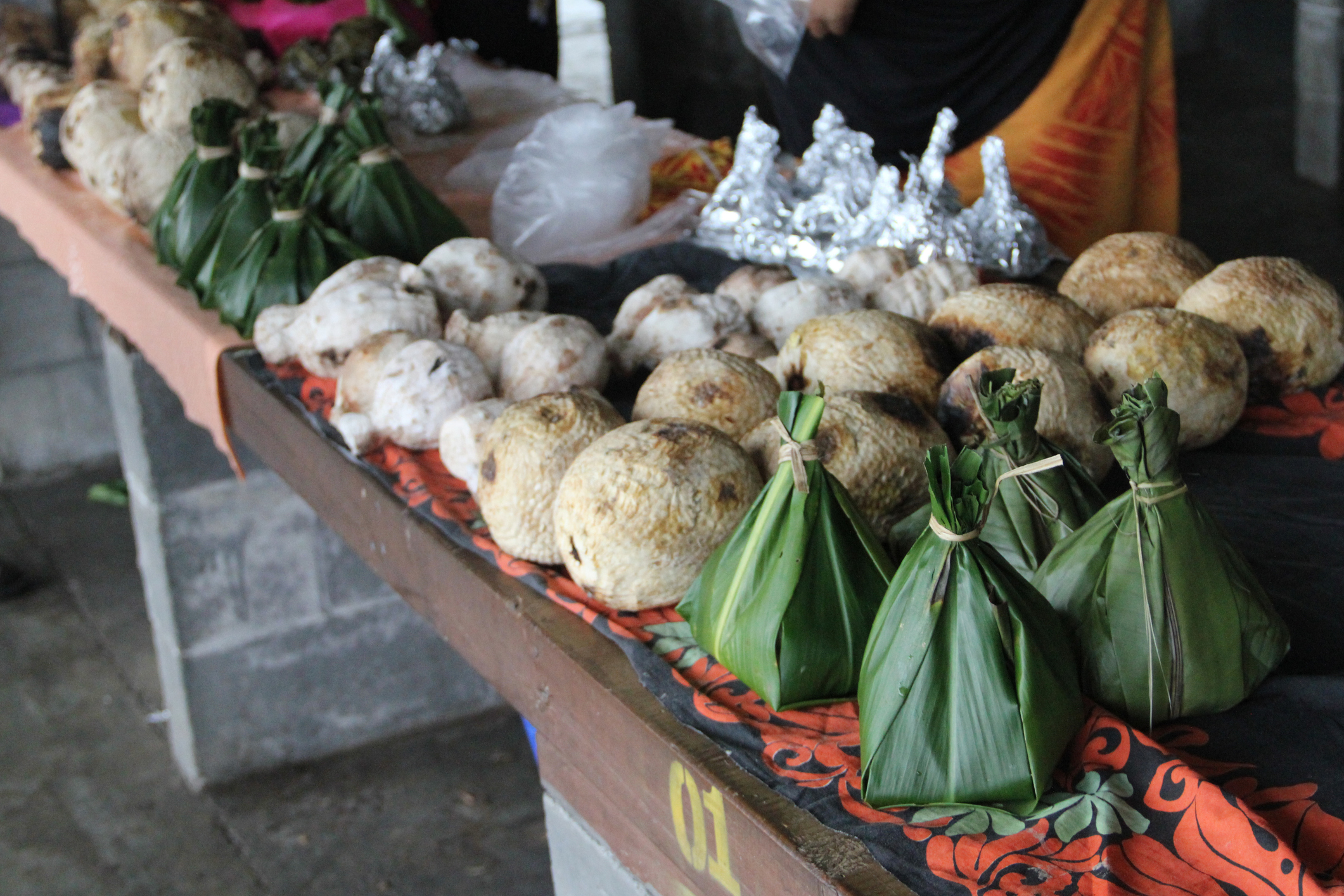 A market stall displays various food items, including round dumplings and bundled parcels wrapped in green leaves.