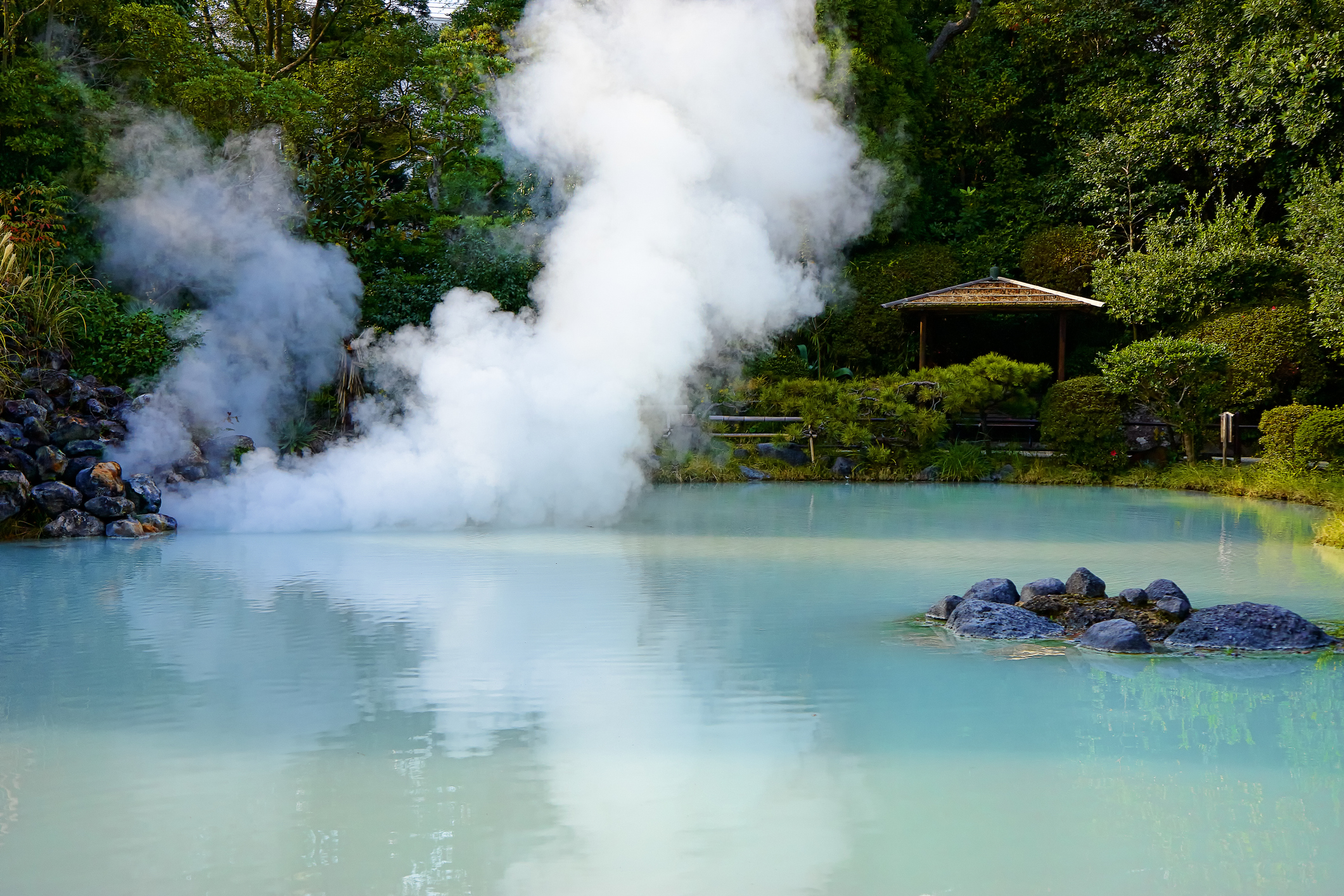 A serene hot spring with light blue water and steam rising, surrounded by lush greenery and a gazebo in the background.