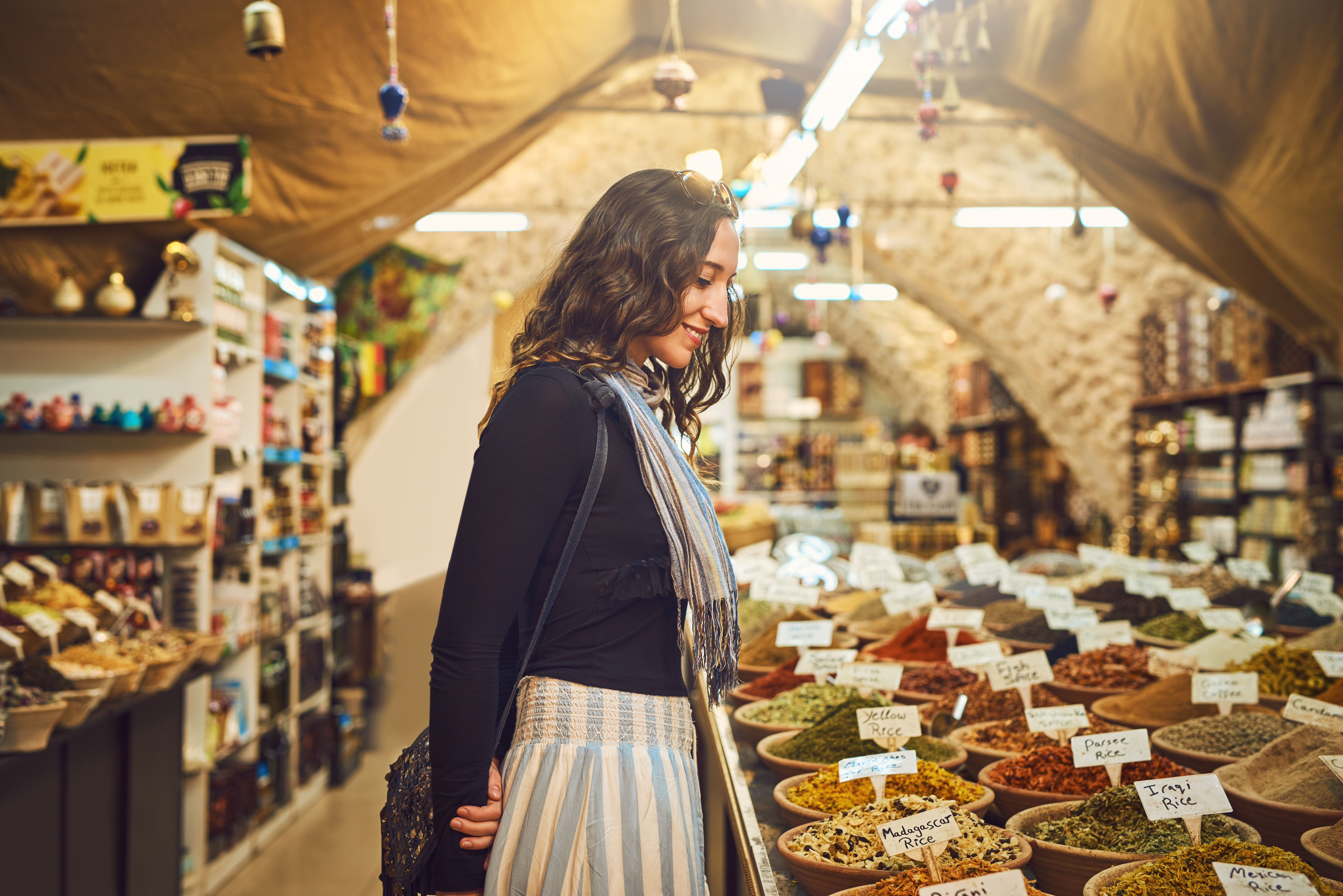A woman stands in a market, admiring colorful spice displays in bowls, surrounded by shelves filled with various products.