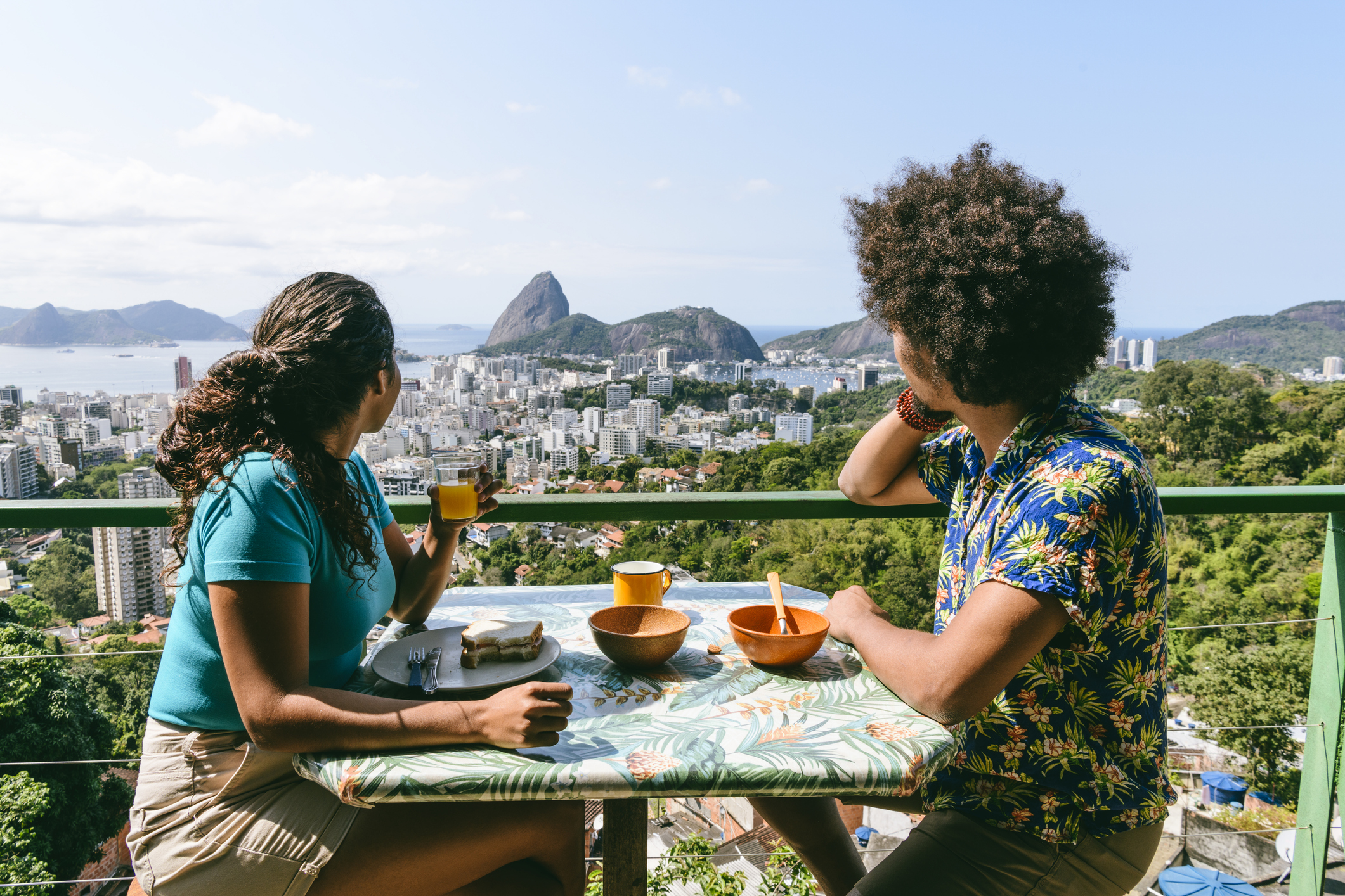 Two people sit at a table with bowls and drinks, overlooking a city and mountainous landscape.