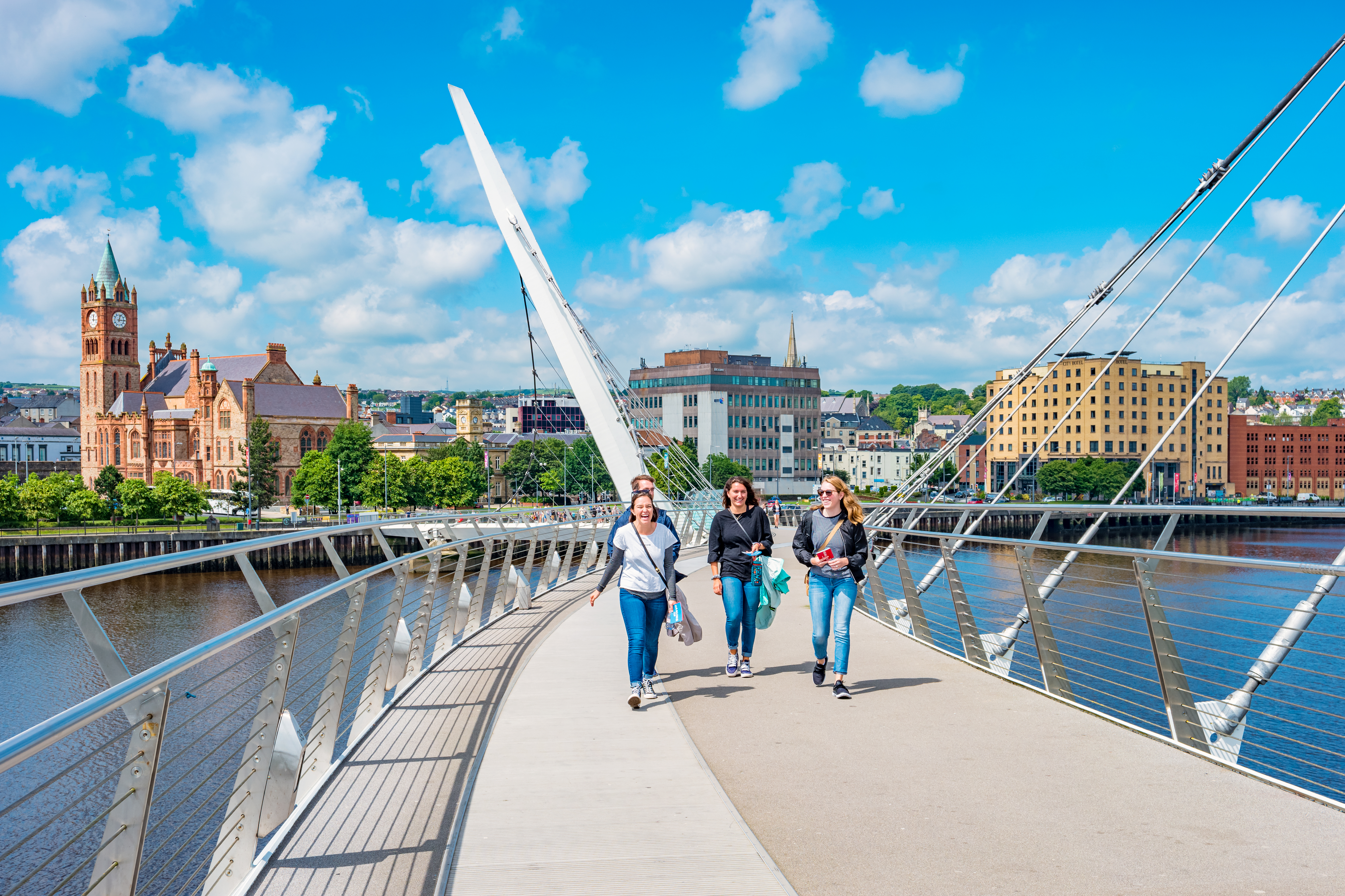 Four people walk across a bridge with a city skyline and blue sky in the background.