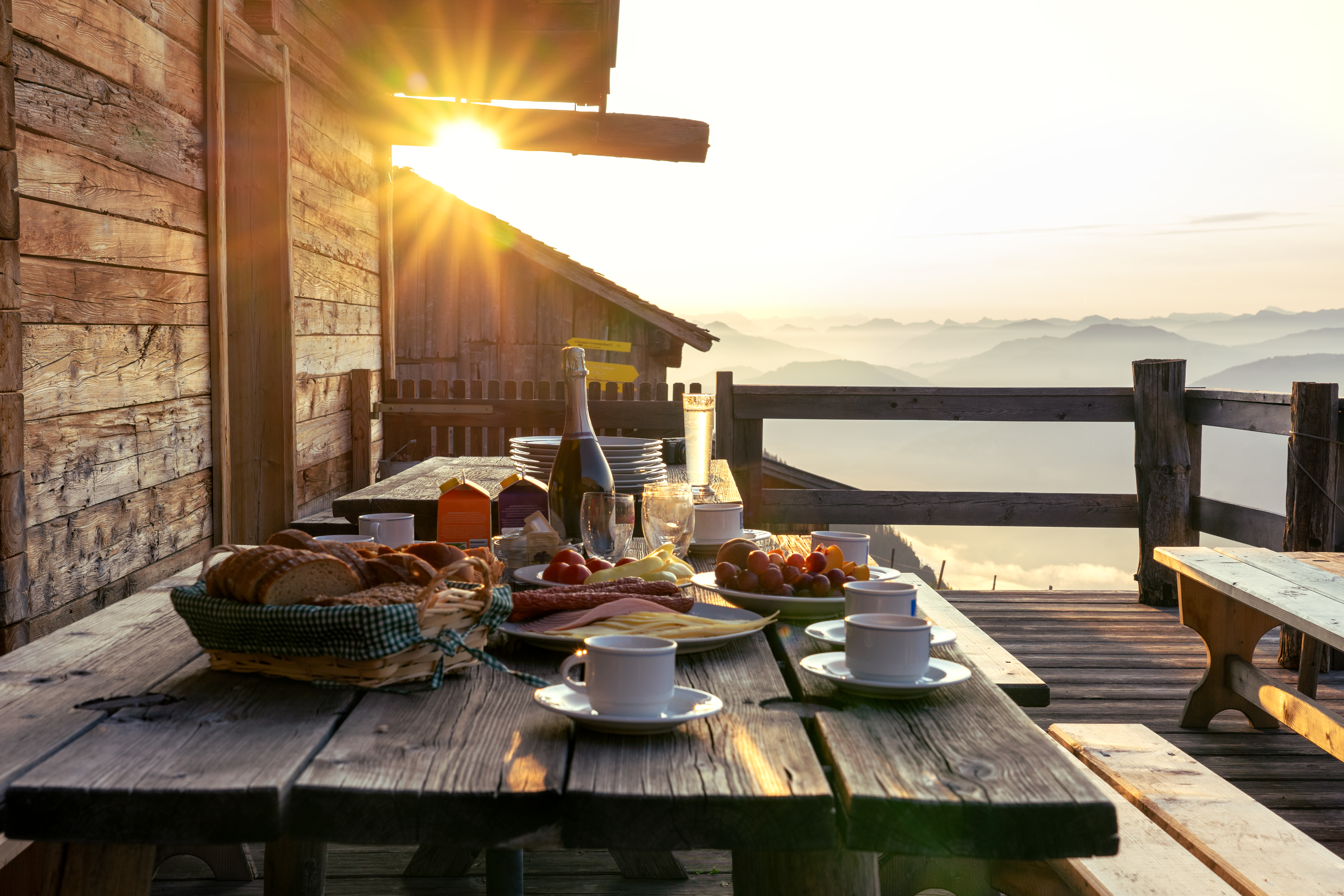 Breakfast spread on a wooden table with coffee, pastries, and fruit, illuminated by sunrise over misty mountains.