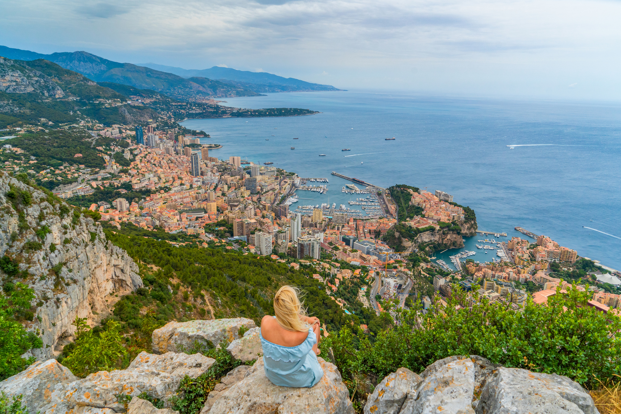 A woman sits on rocks overlooking a coastal city, with buildings and boats nestled along the shoreline and hills in the background.