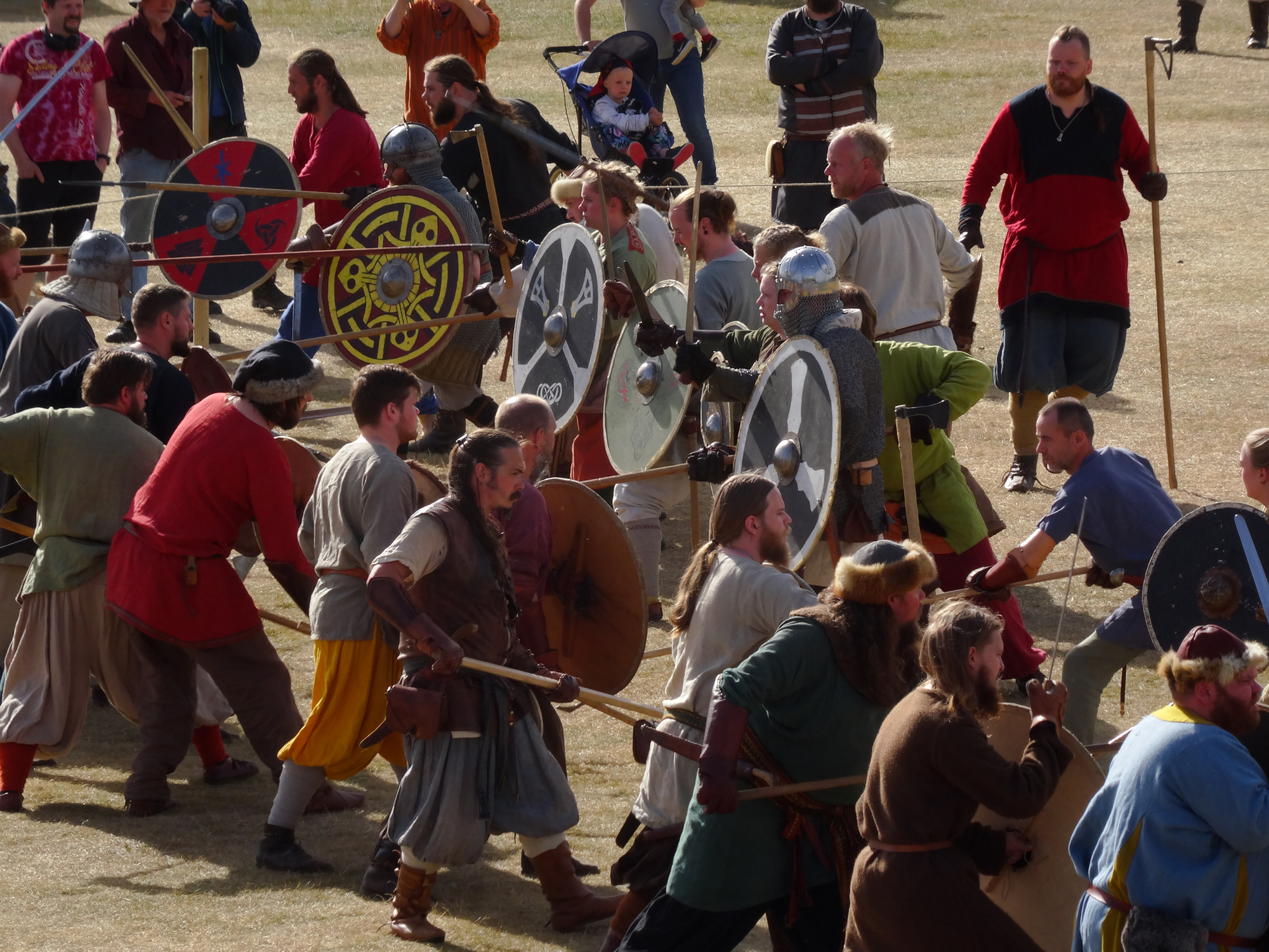 A reenactment scene with people in medieval costumes wielding shields and weapons in a grassy area.