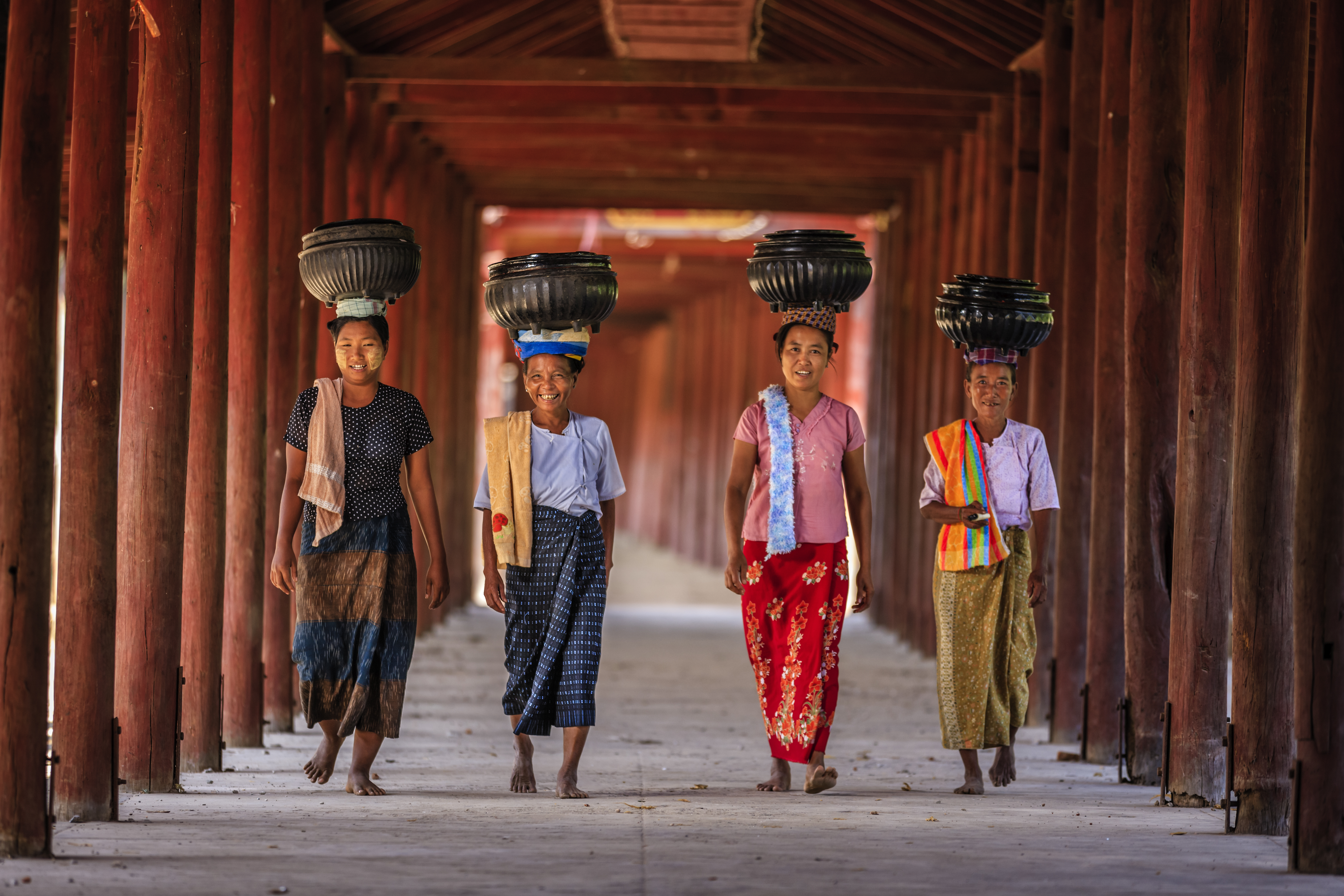 Four girls walk confidently under a wooden archway, each balancing a container on their heads.