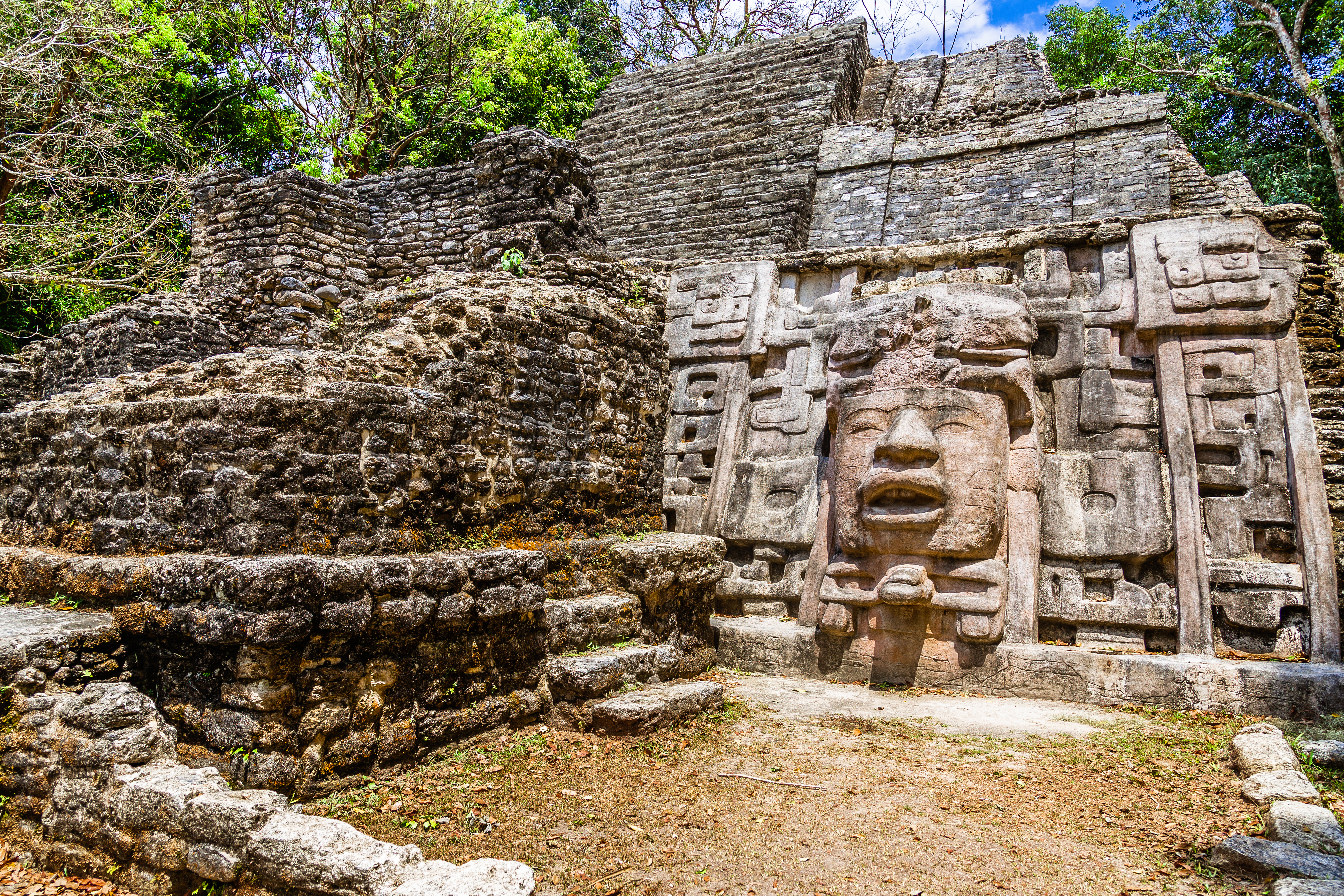 Ancient stone ruins with a carved face, surrounded by greenery and blue sky.