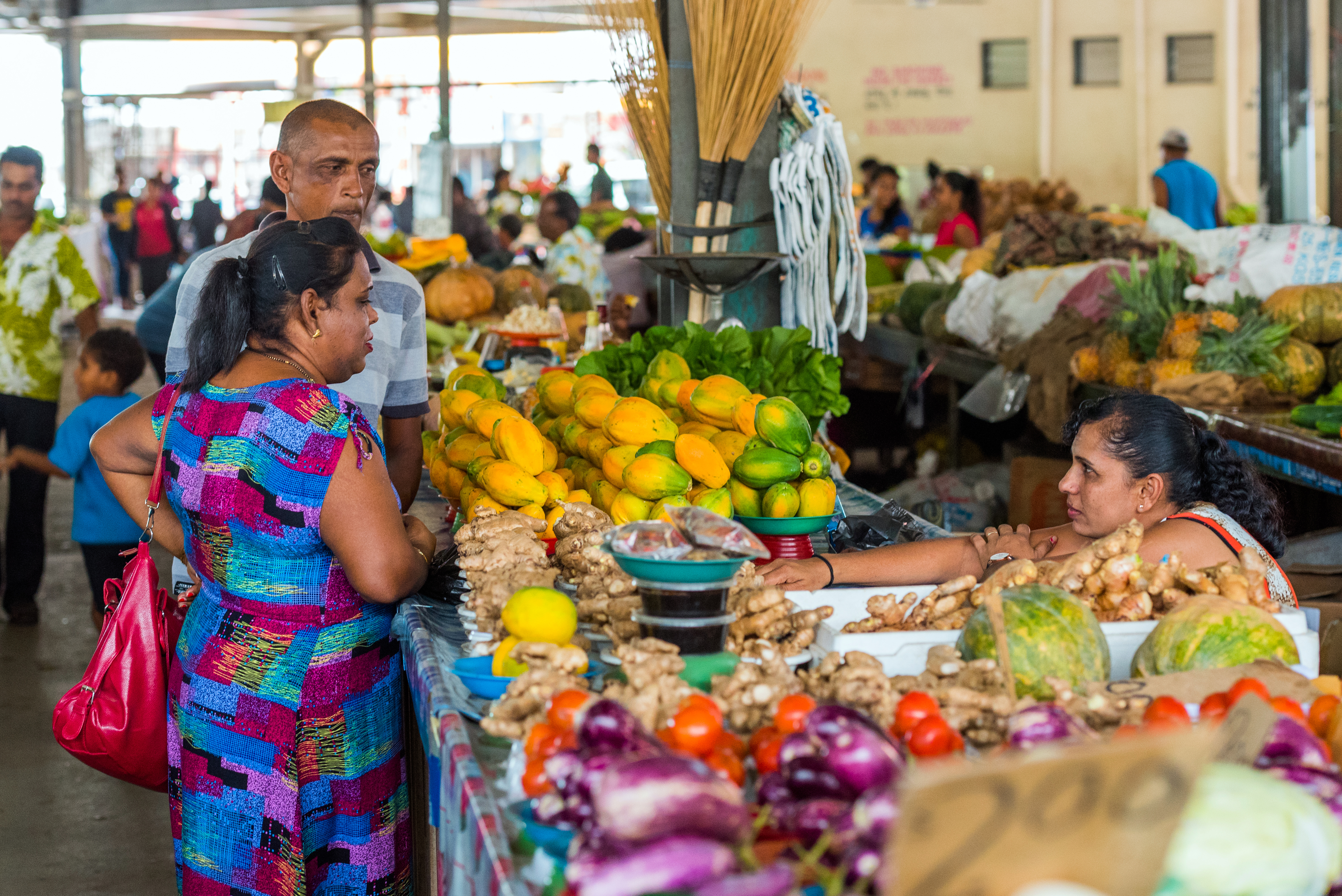 Two women and a man shop at a vibrant market, surrounded by fresh fruits and vegetables in a lively atmosphere.