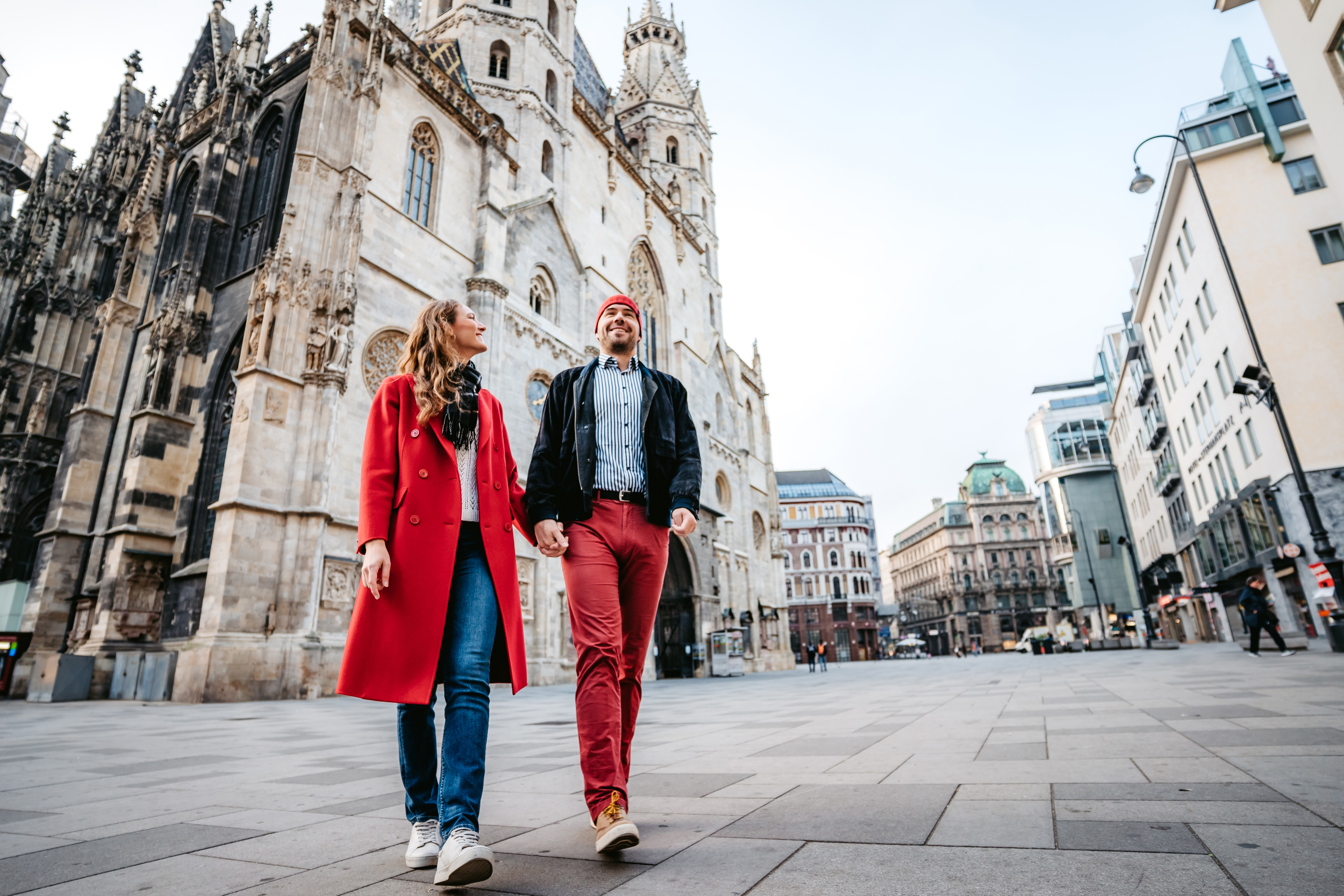 A couple walks hand in hand through a city square, with historical architecture in the background.