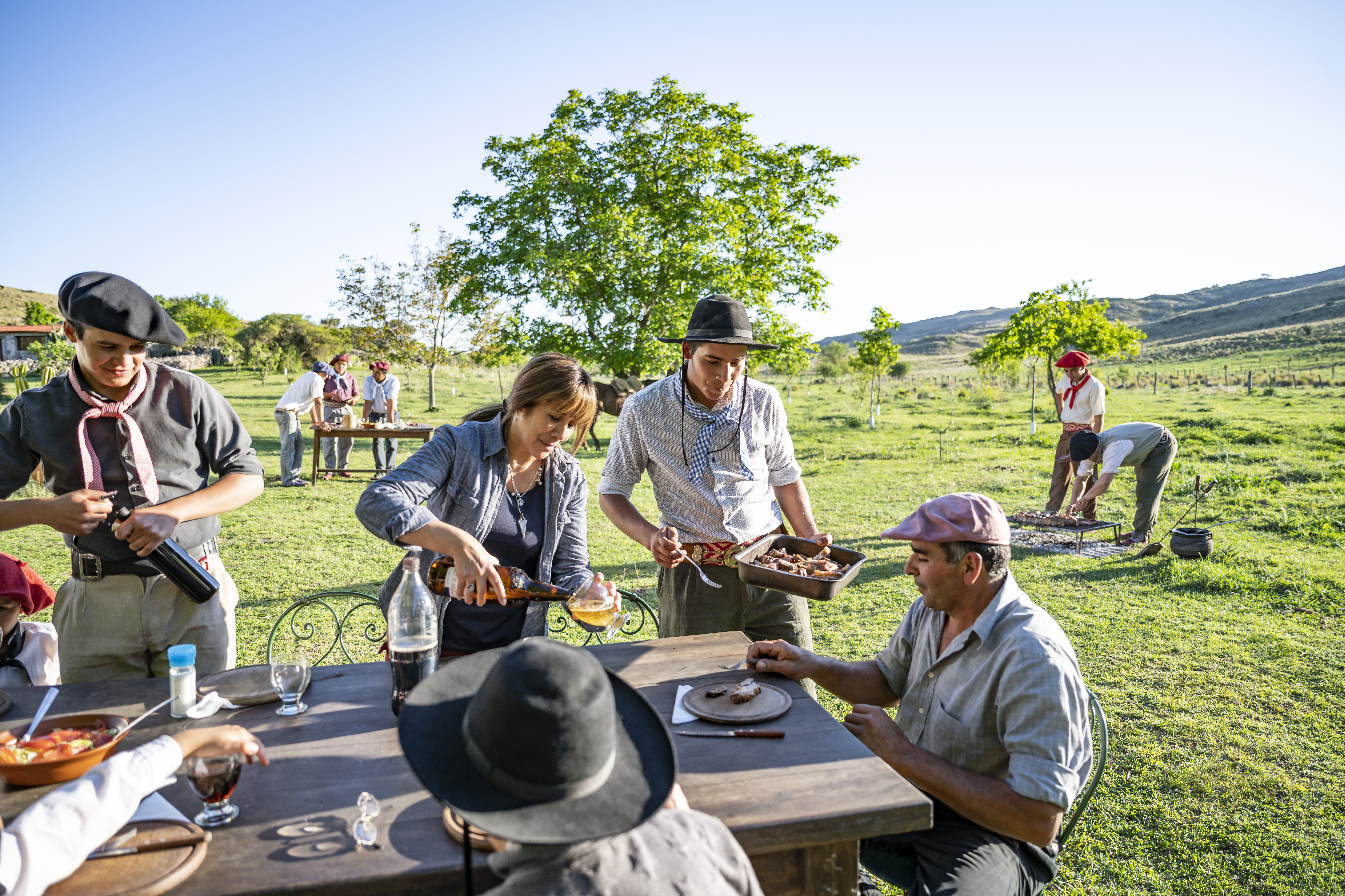 A gathering in a rural setting with people serving food at a table under a clear blue sky and trees.