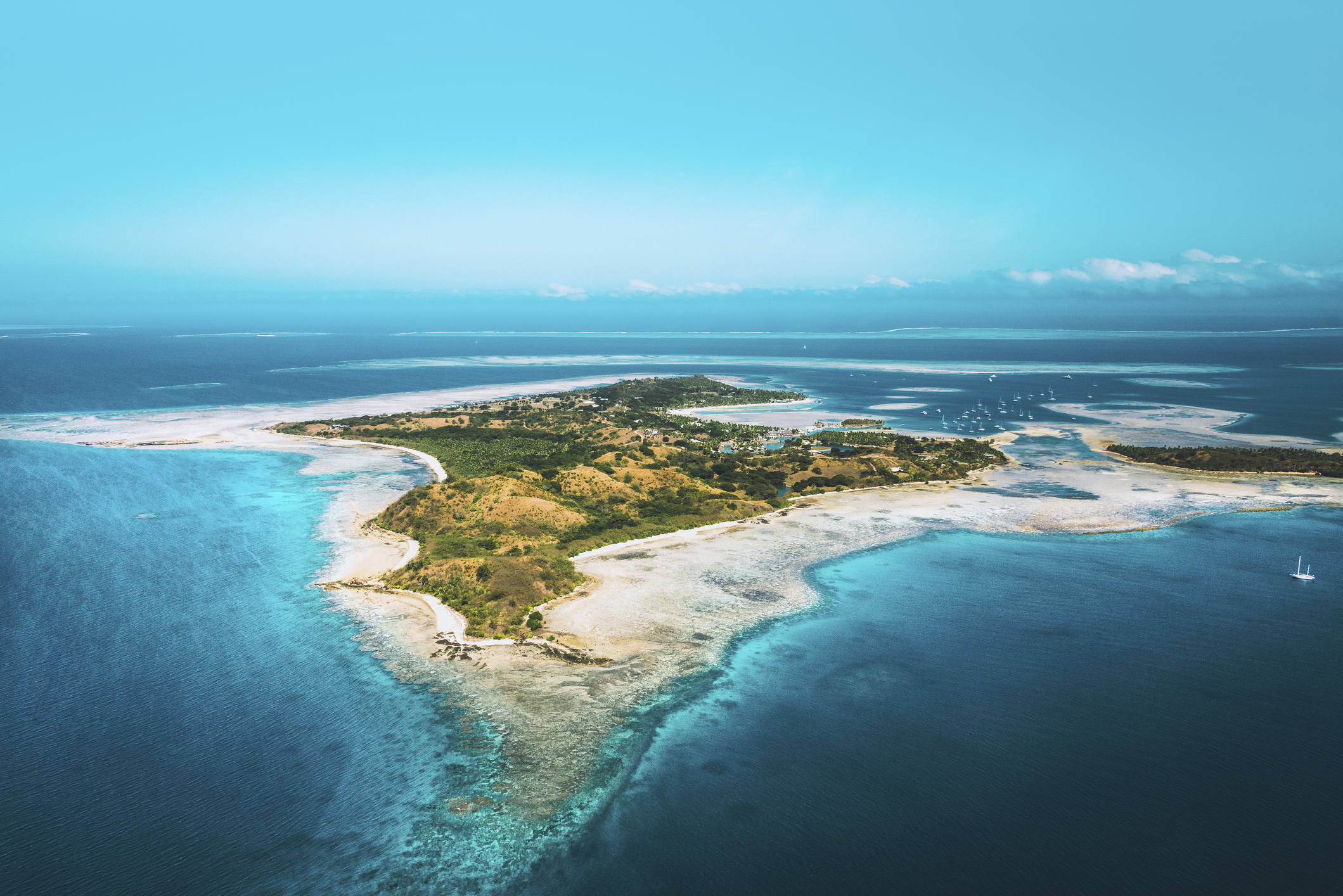 Aerial view of a small, lush island surrounded by turquoise water and coral reefs under a clear blue sky.