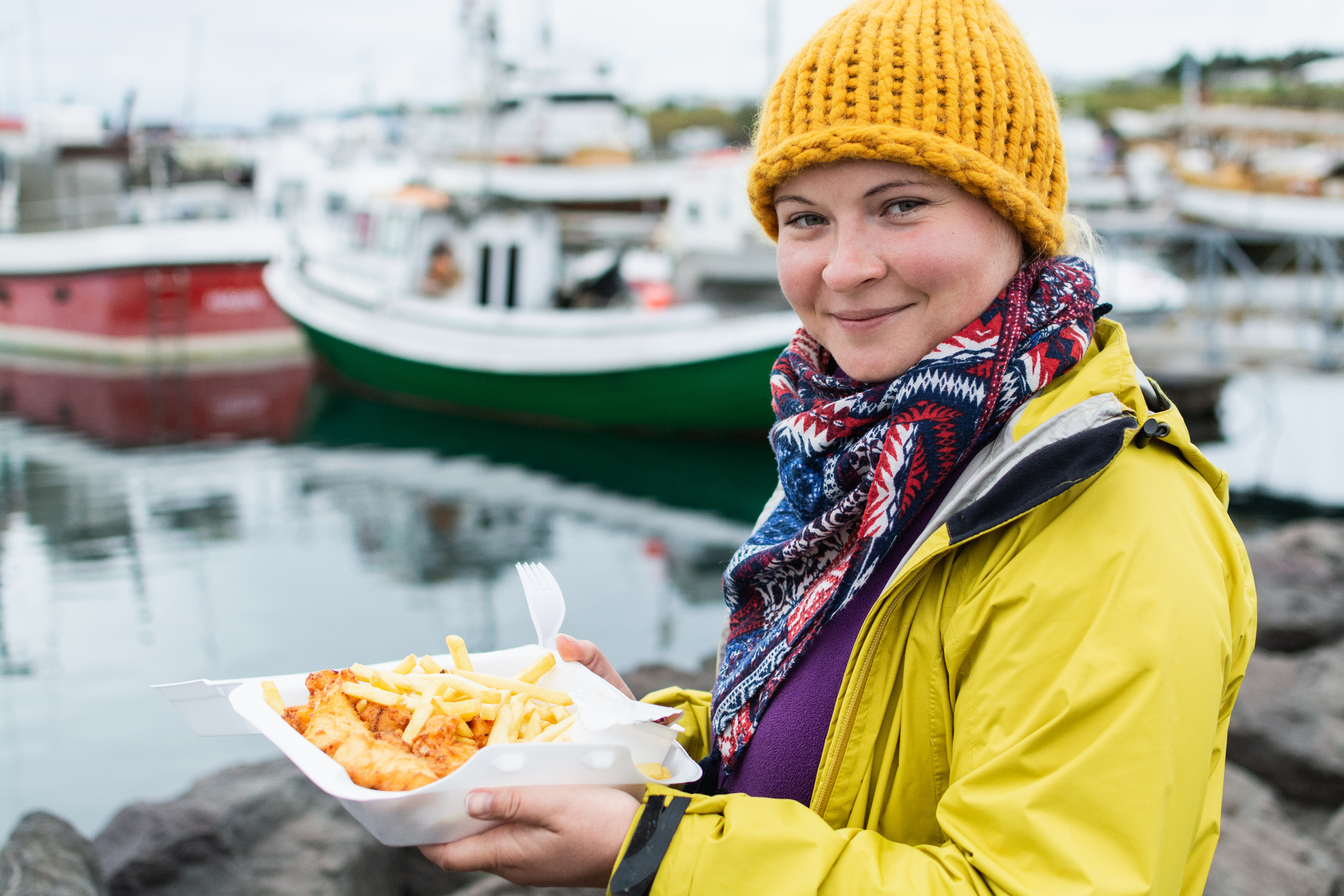 A smiling person in a yellow jacket and knit hat holds a tray of fish and chips by the water, with boats in the background.