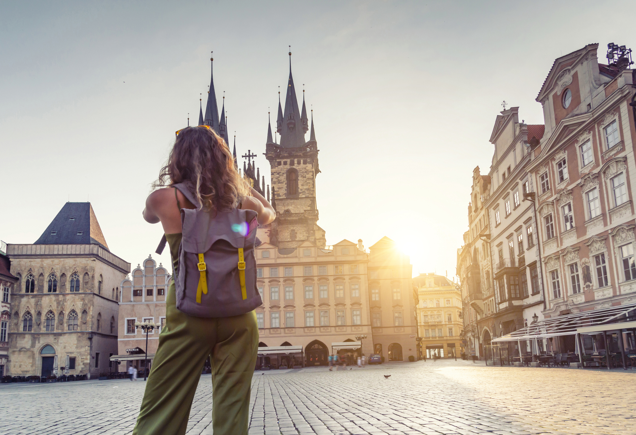 A person with a backpack stands in an empty square, gazing at historic buildings under a golden sunset.